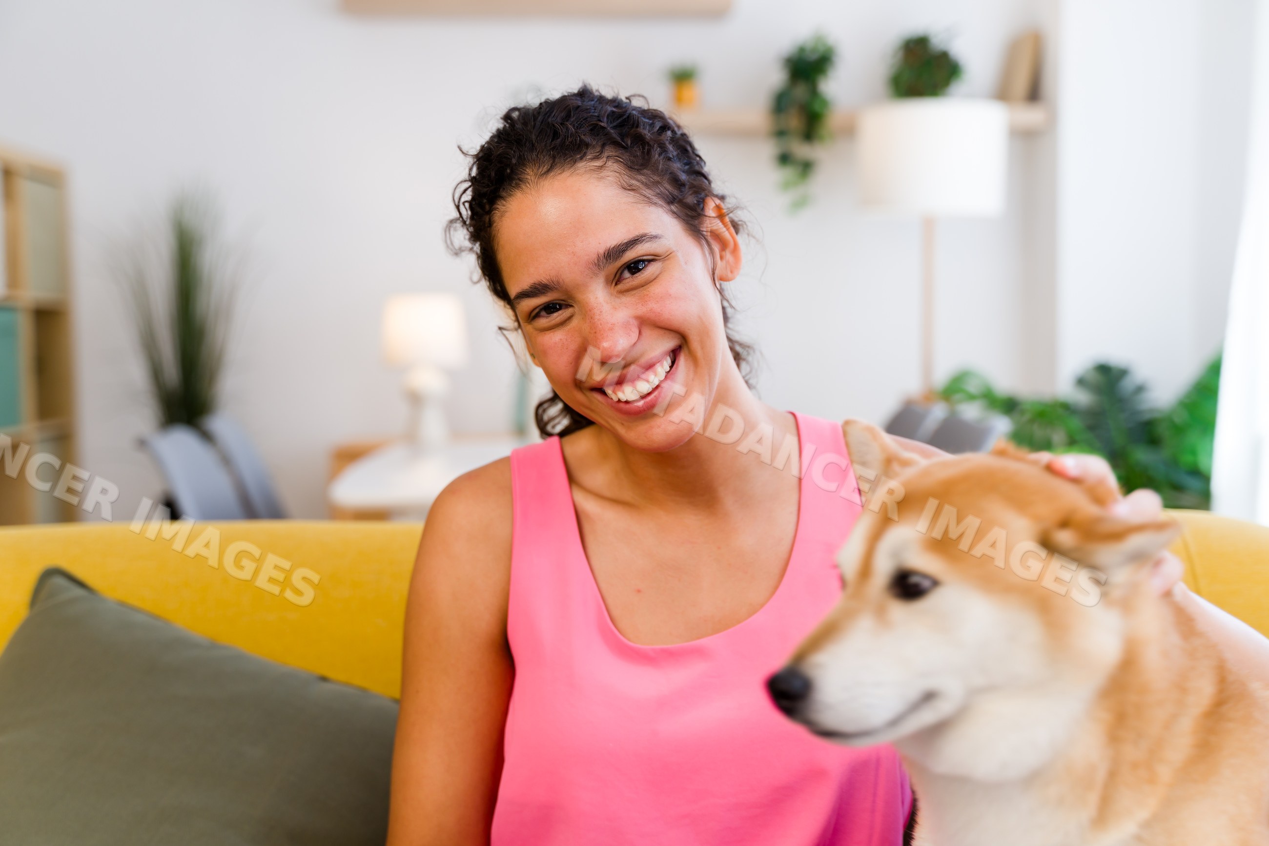 A Hispanic woman happily pets her dog on a yellow sofa