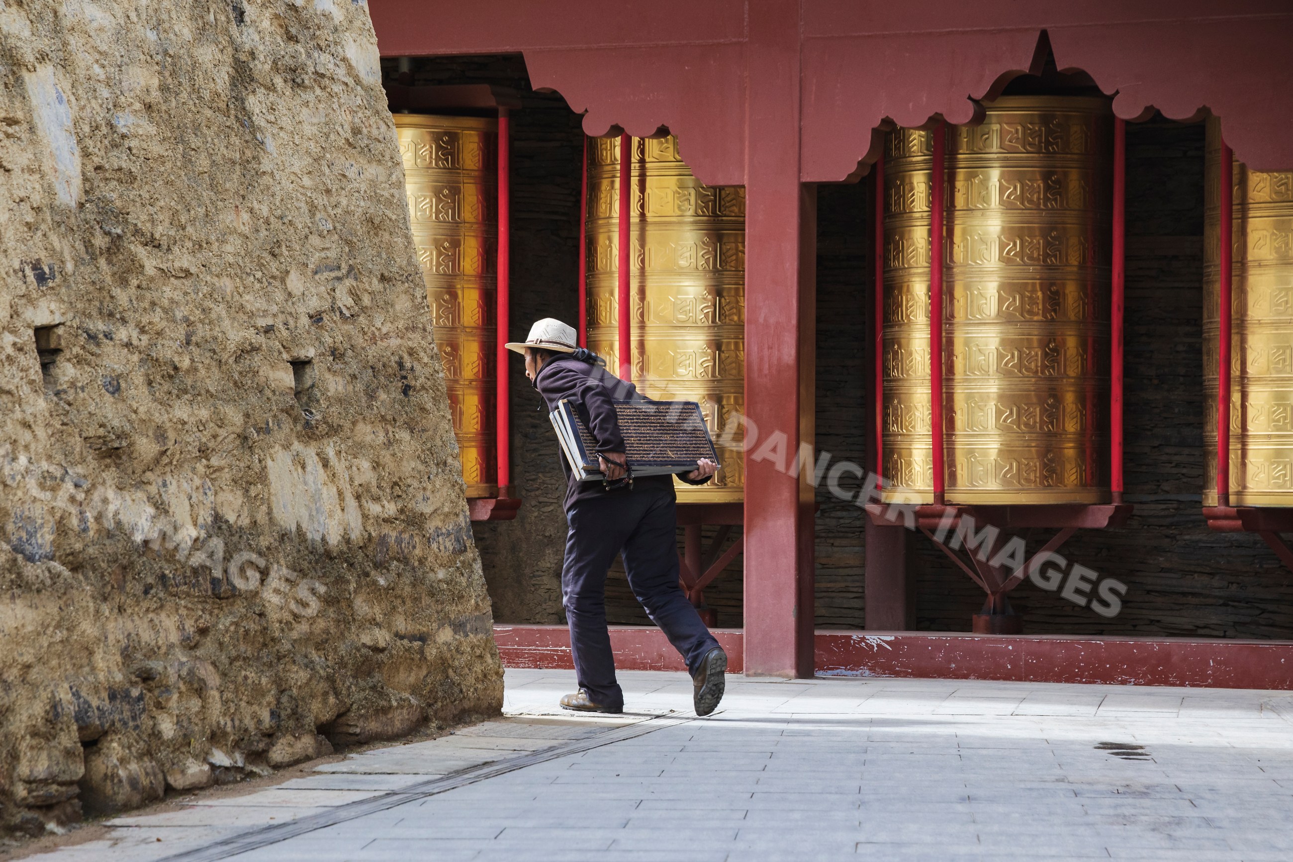 Tibetan man carrying inscribed tablet by Mani Wall with prayer wheels