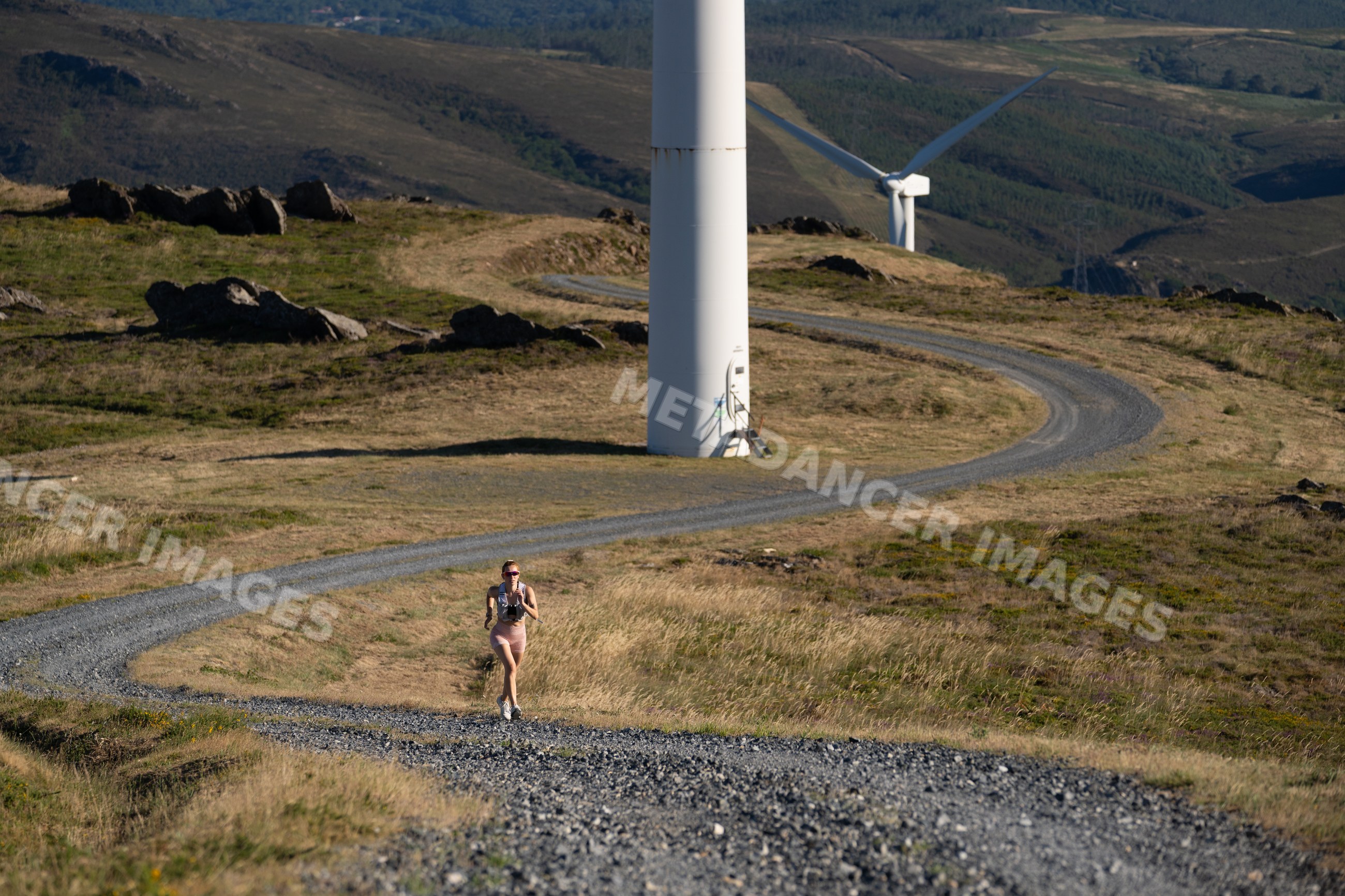 A white woman runs on a winding trail by wind turbines in Galicia