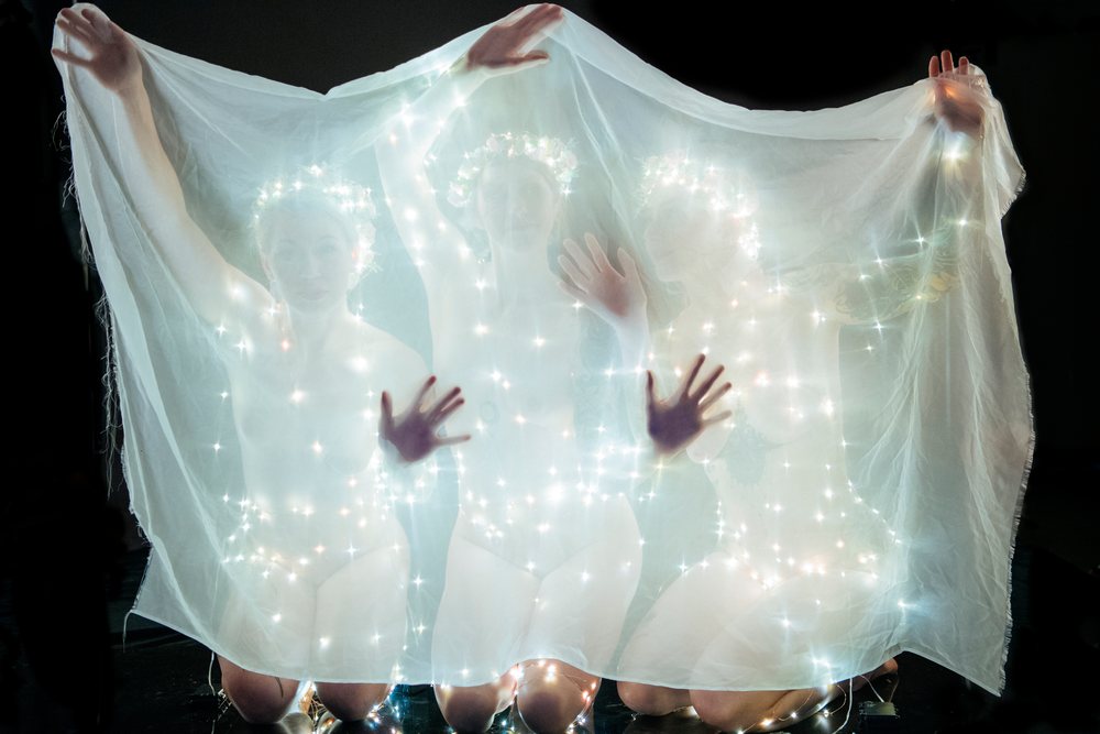 Three white women kneeling behind sheer fabric with glowing string