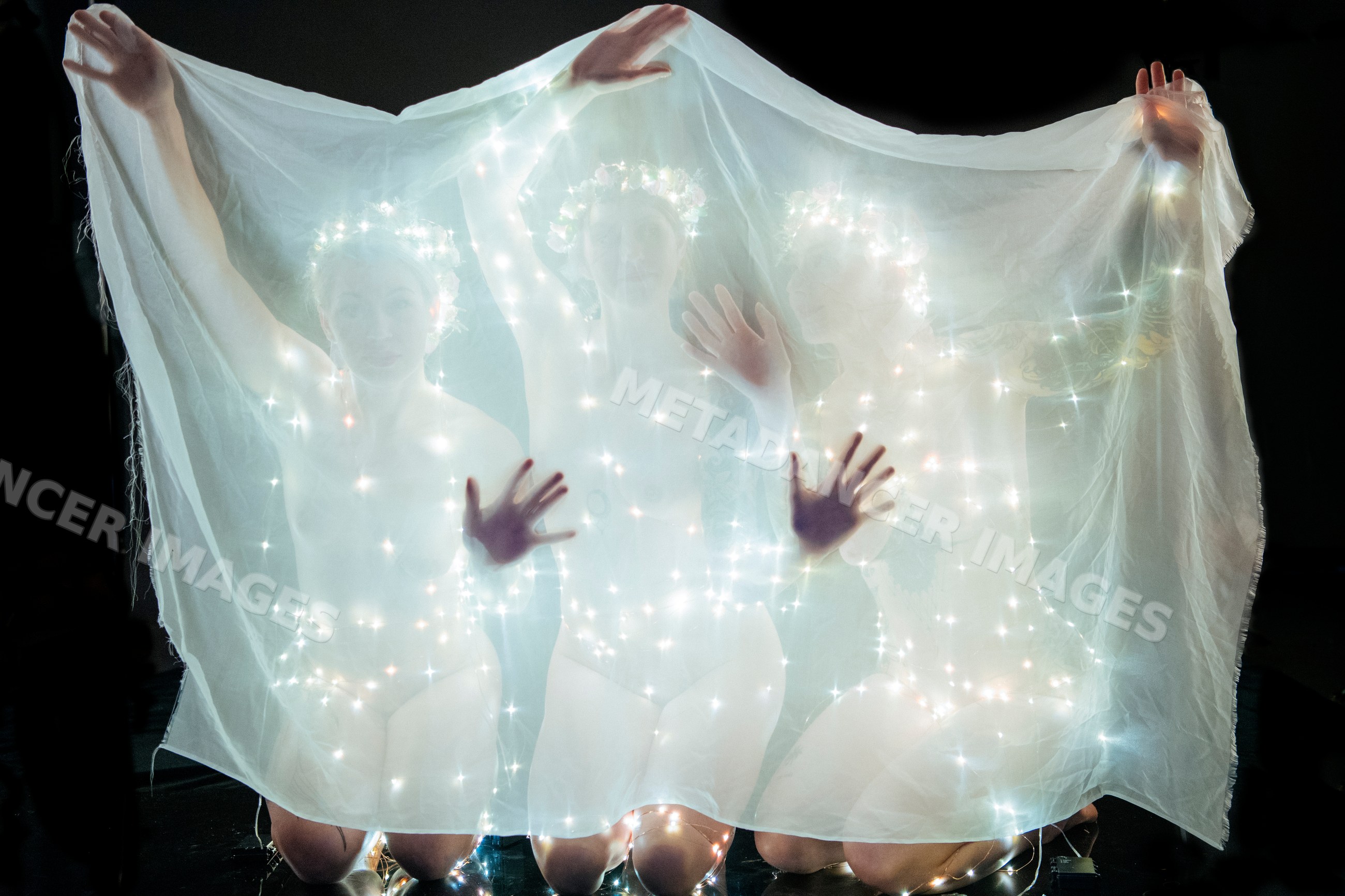 Three white women kneeling behind sheer fabric with glowing string