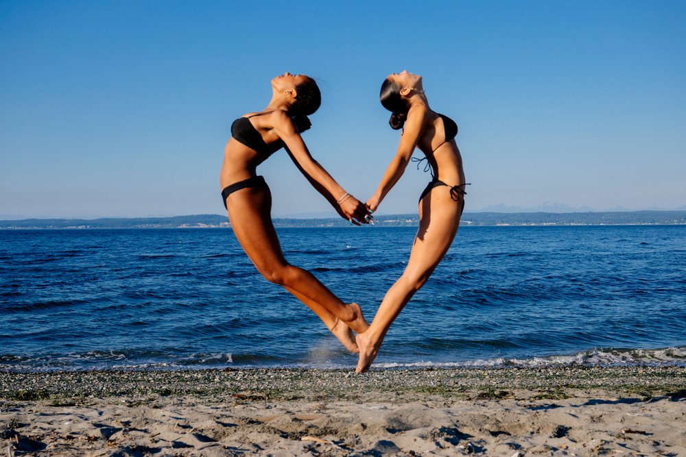 Two women doing acrobatic yoga poses holding hands on a beach