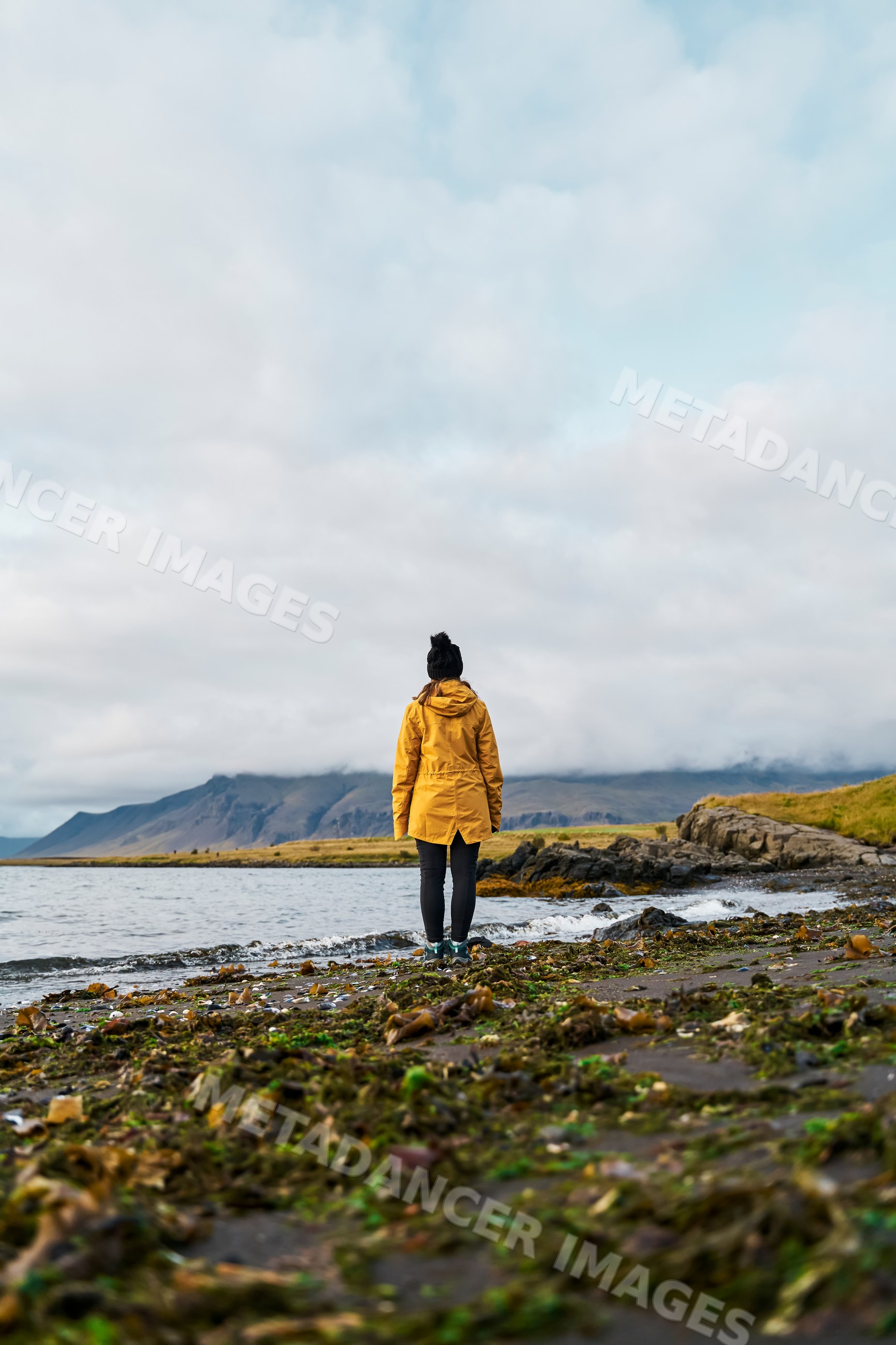 A white woman in a yellow jacket stands on a rocky shore