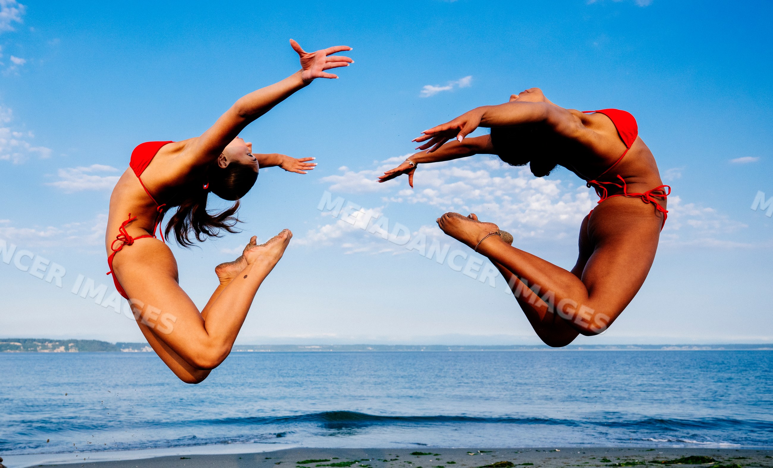 Two women jump in synchrony doing partner acrobatics on a beach
