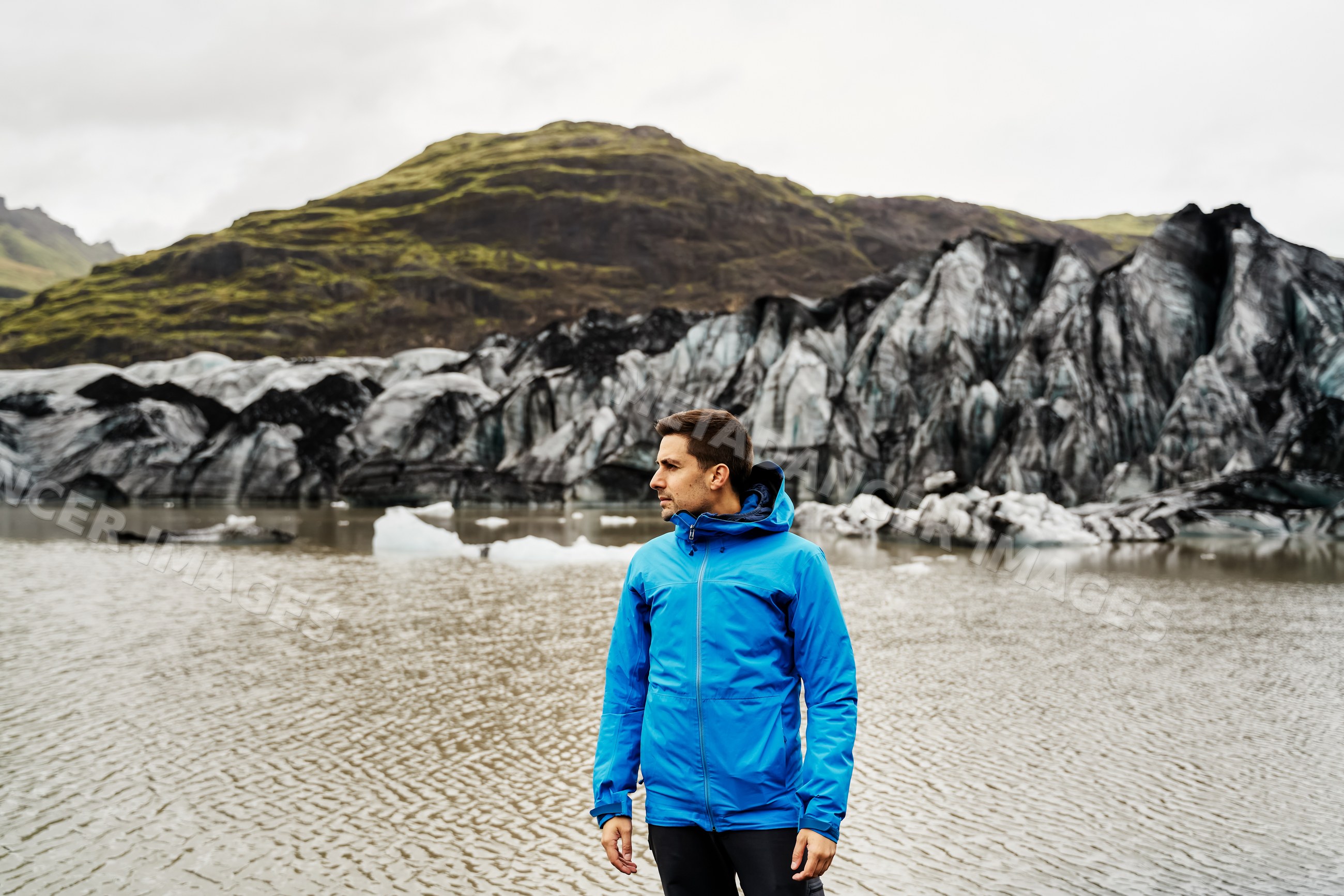 A white man stands by a glacier lake with mountains