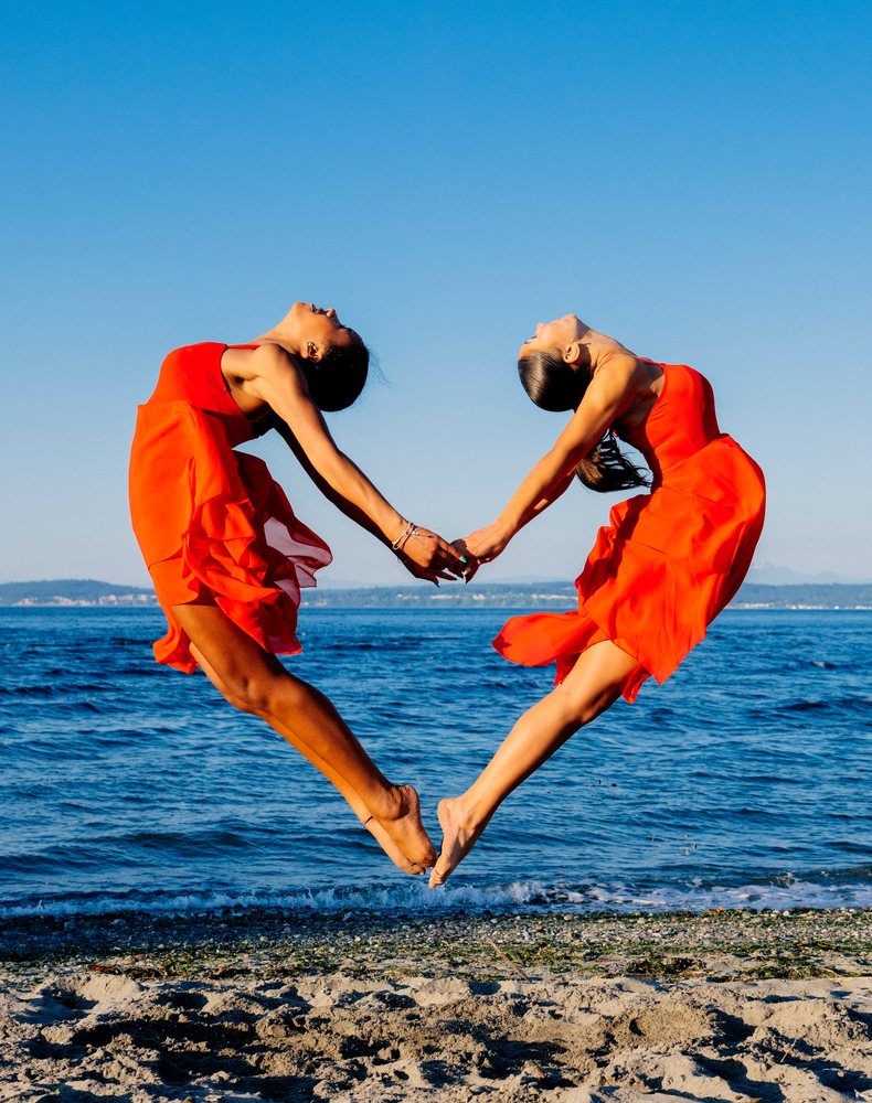Two women in red dresses perform partner acrobatics jumping on a beach