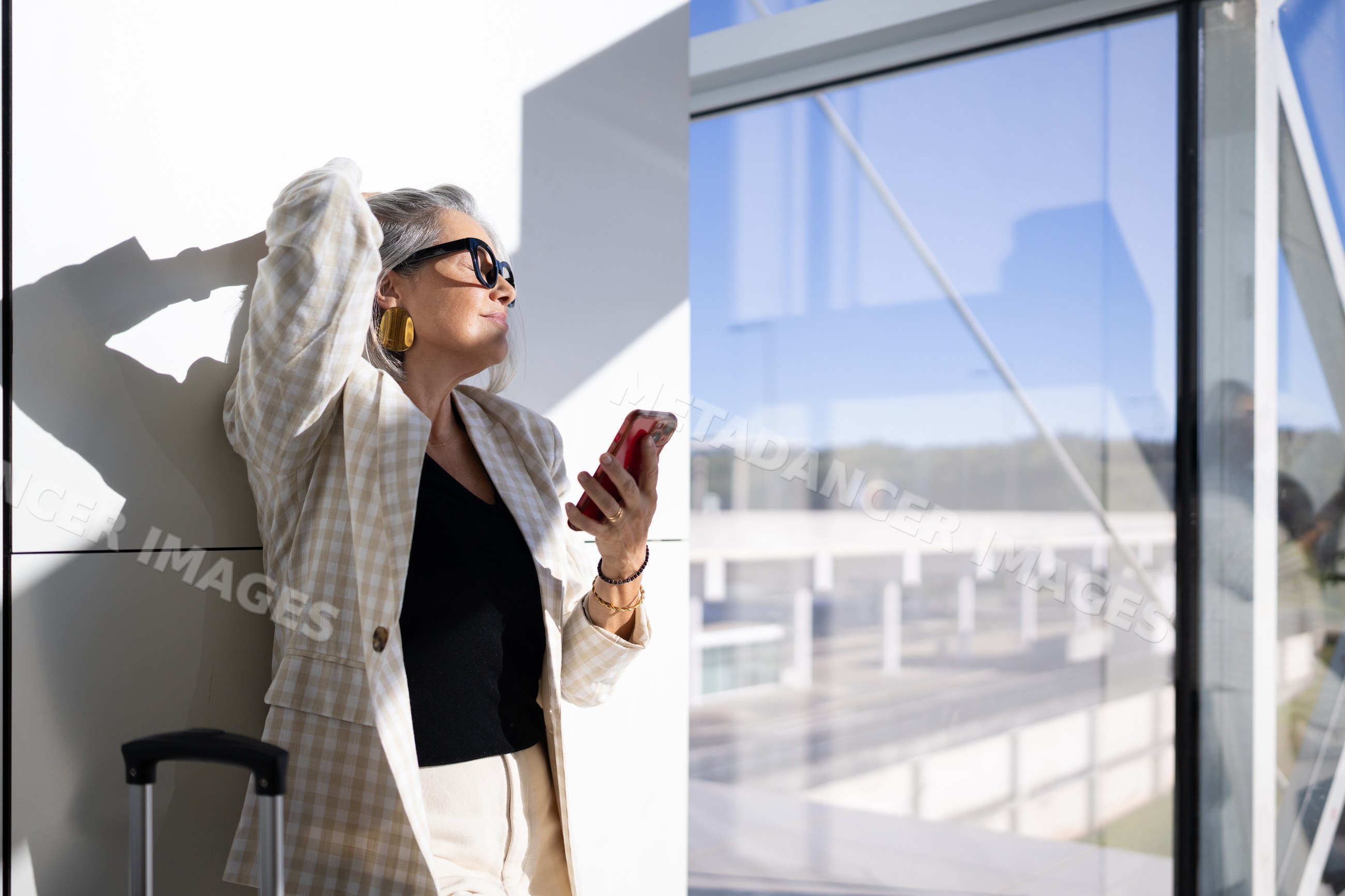 White woman standing indoors near window with mobile phone in sunlight
