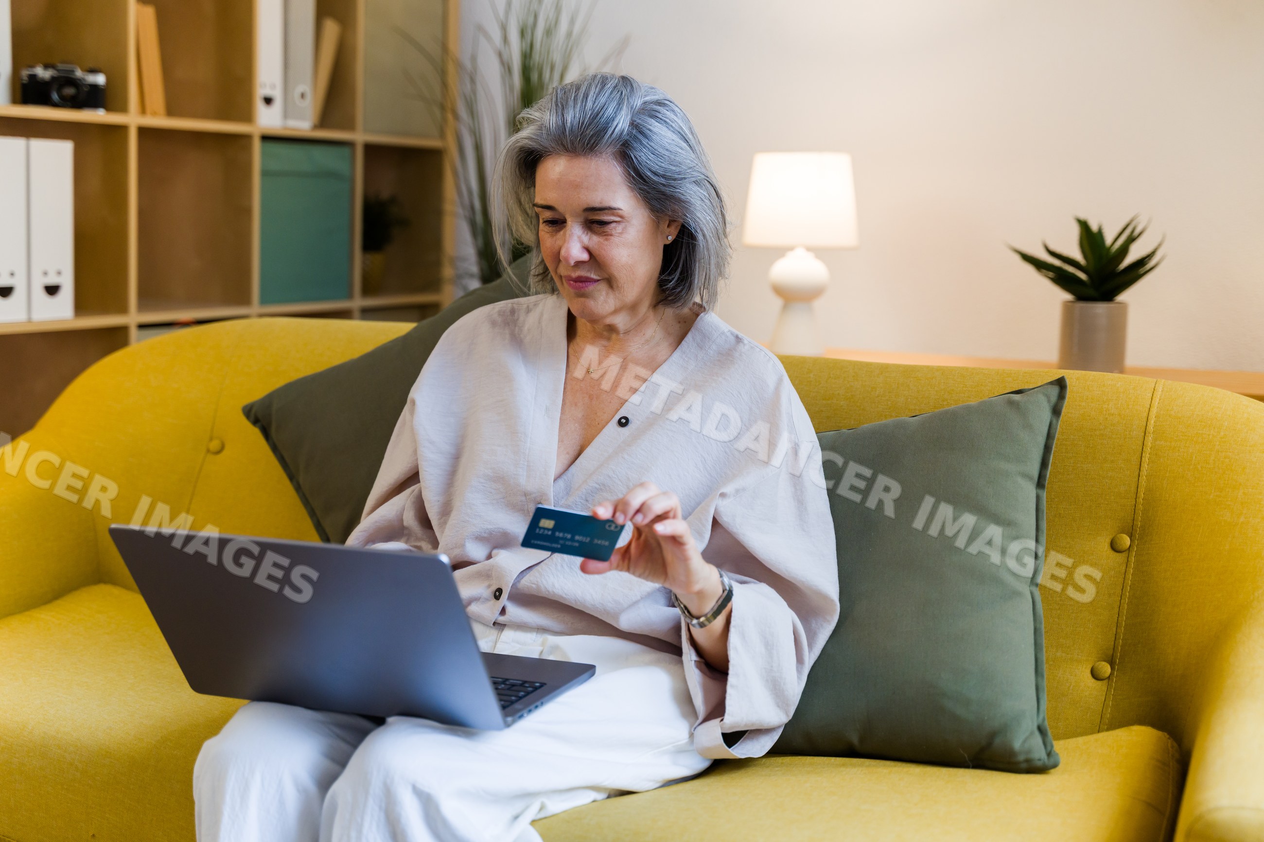 White woman with credit card using laptop on yellow sofa indoors