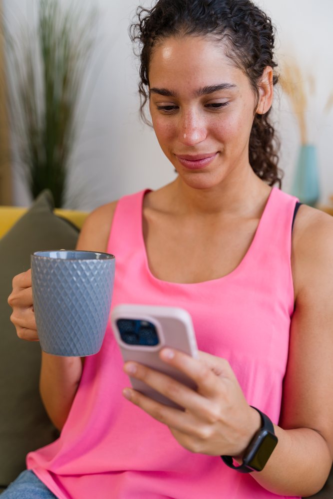 A Hispanic woman relaxes on a couch using her smartphone and drinking