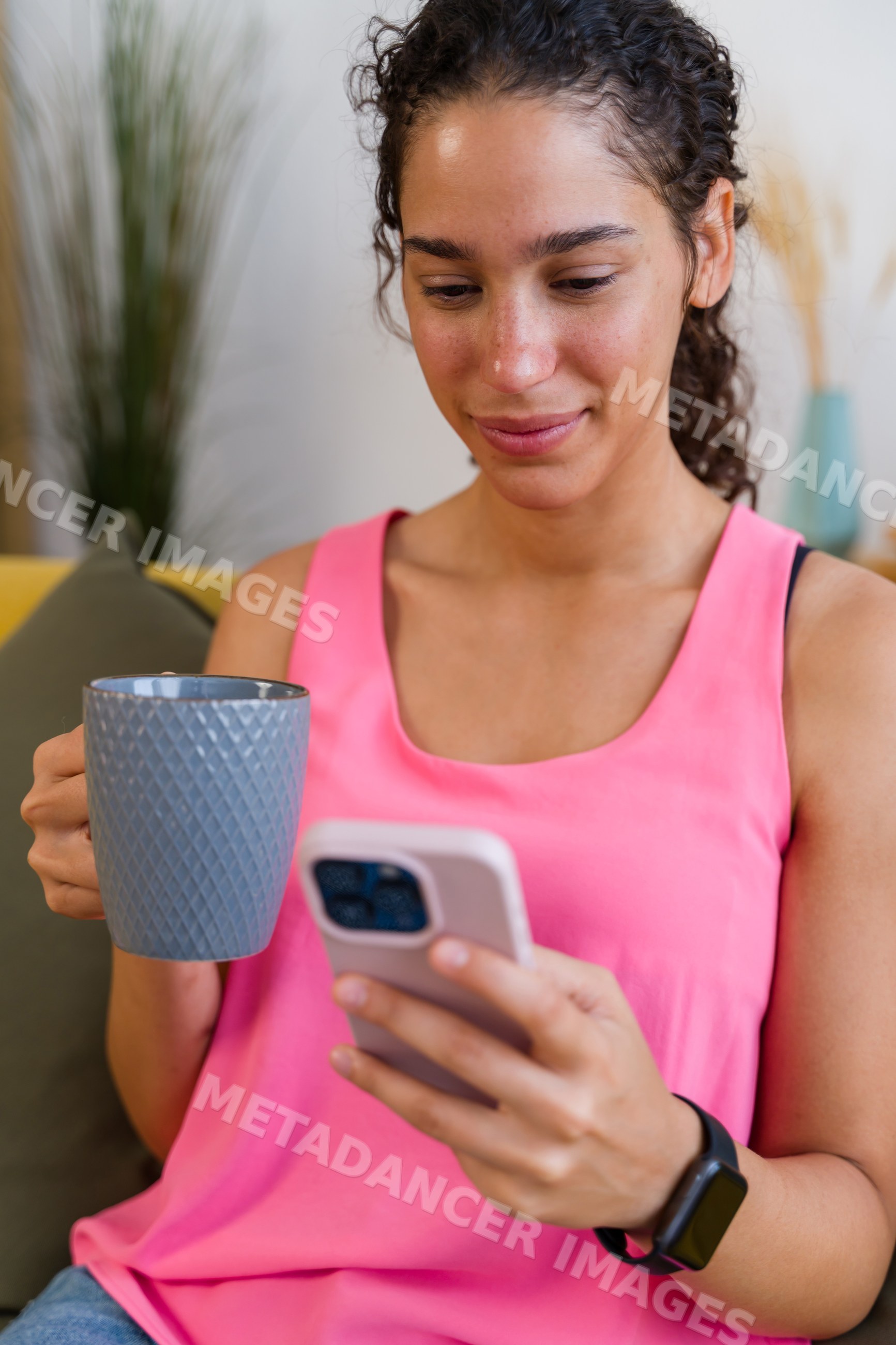 A Hispanic woman relaxes on a couch using her smartphone and drinking