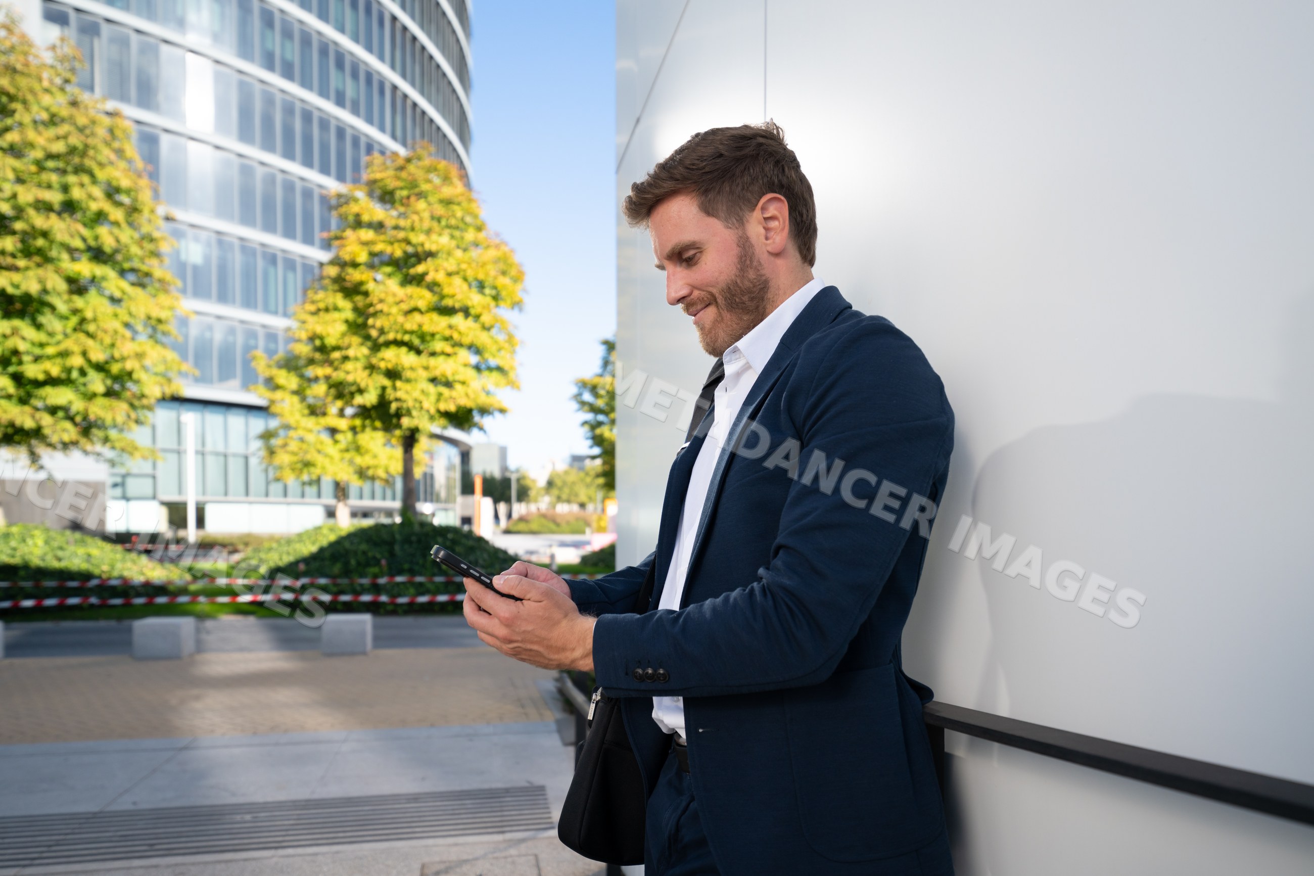 White man using smartphone leaning on wall outside modern office