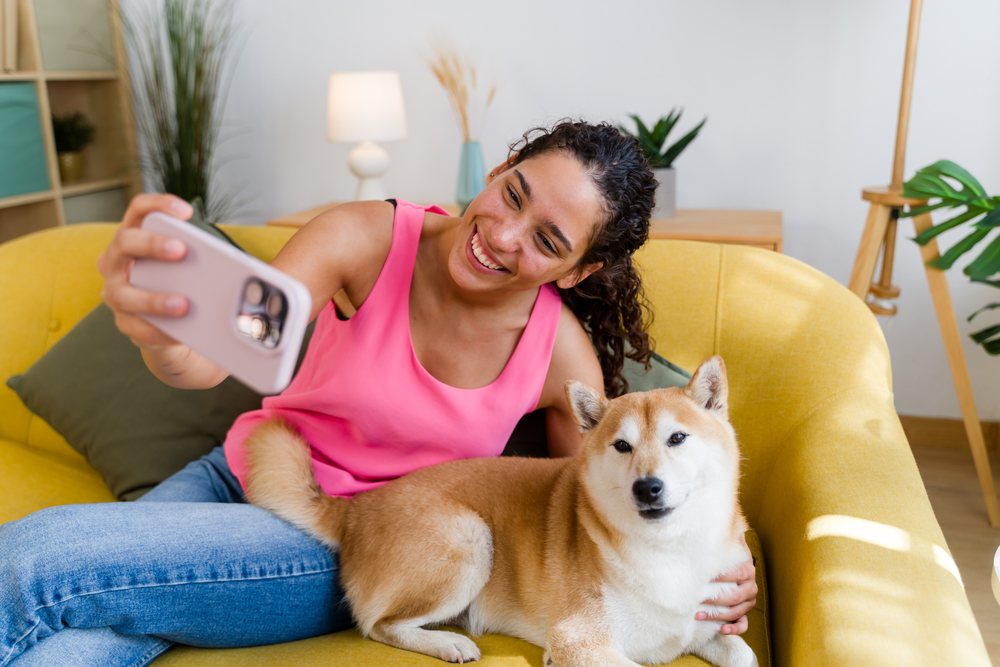 A Hispanic woman takes a selfie with her dog on a yellow couch