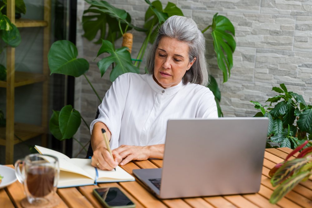Mature white woman writing in notebook at wooden table with laptop