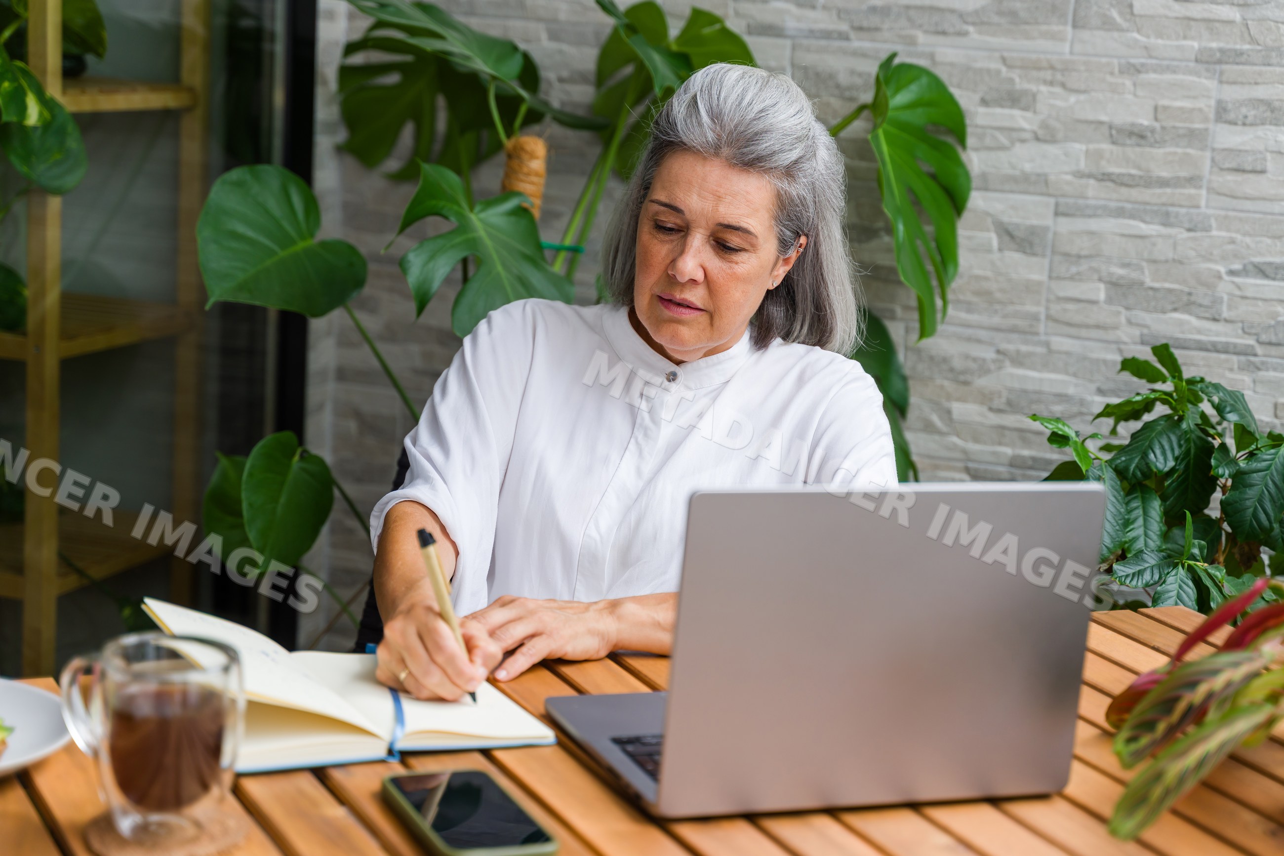 Mature white woman writing in notebook at wooden table with laptop