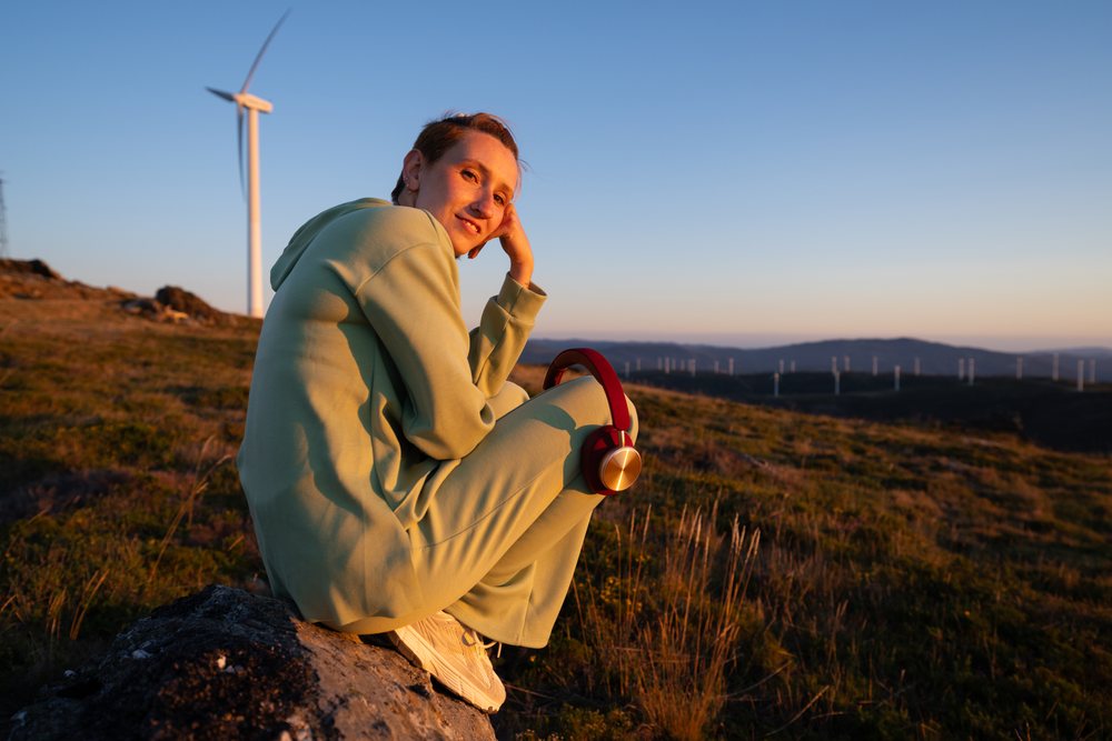 Young white woman sitting with headphones near wind turbines at sunset