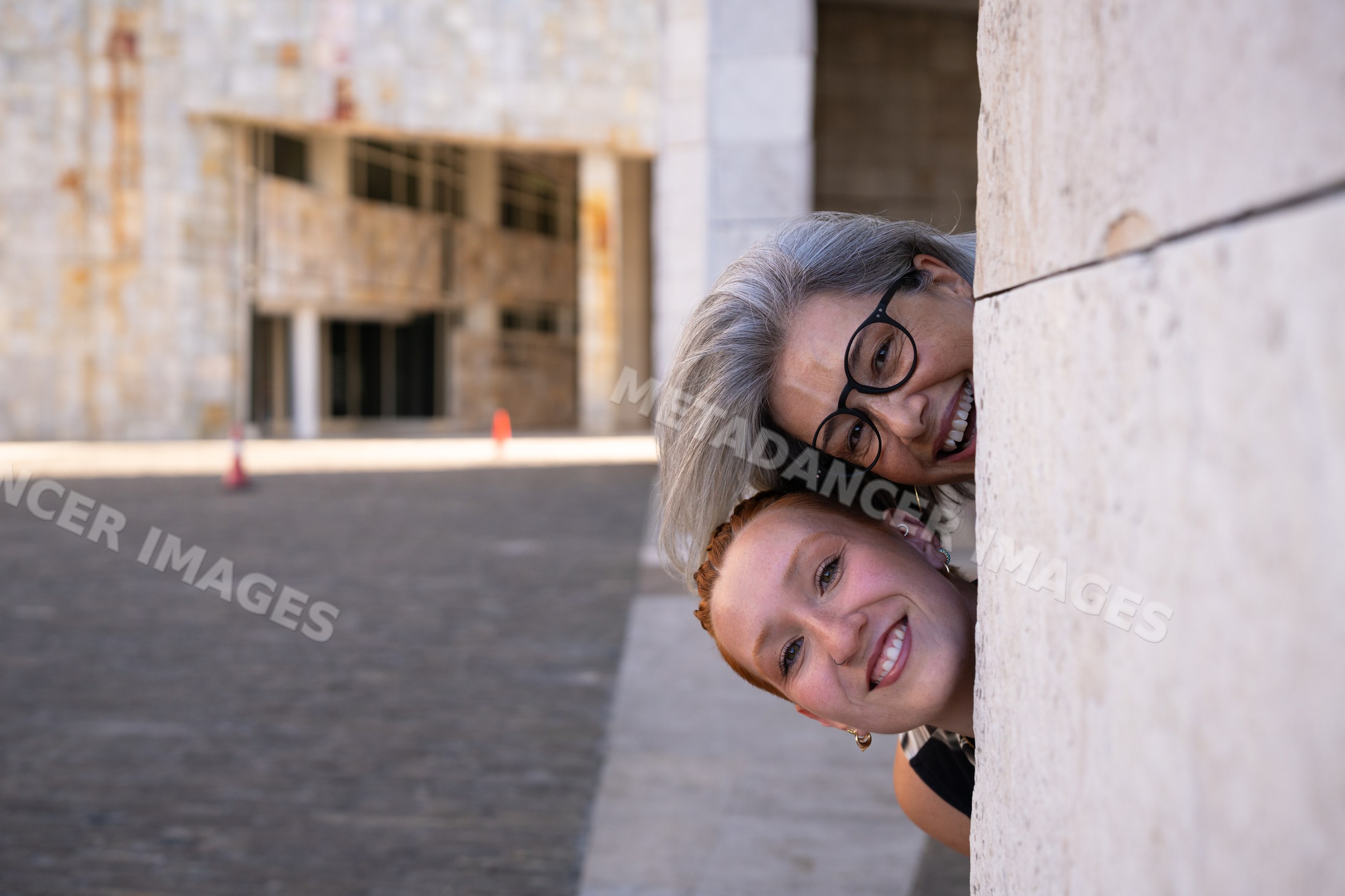 Two white women laughing and peeking around a stone wall outdoors