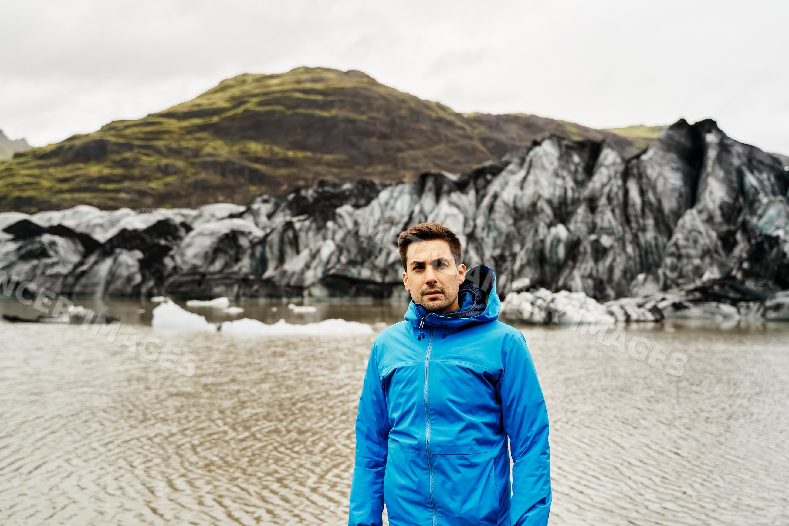 A white man in a blue jacket stands by a glacier lake and mountains