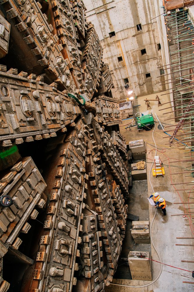 Two white workers review plans by a tunnel boring machine