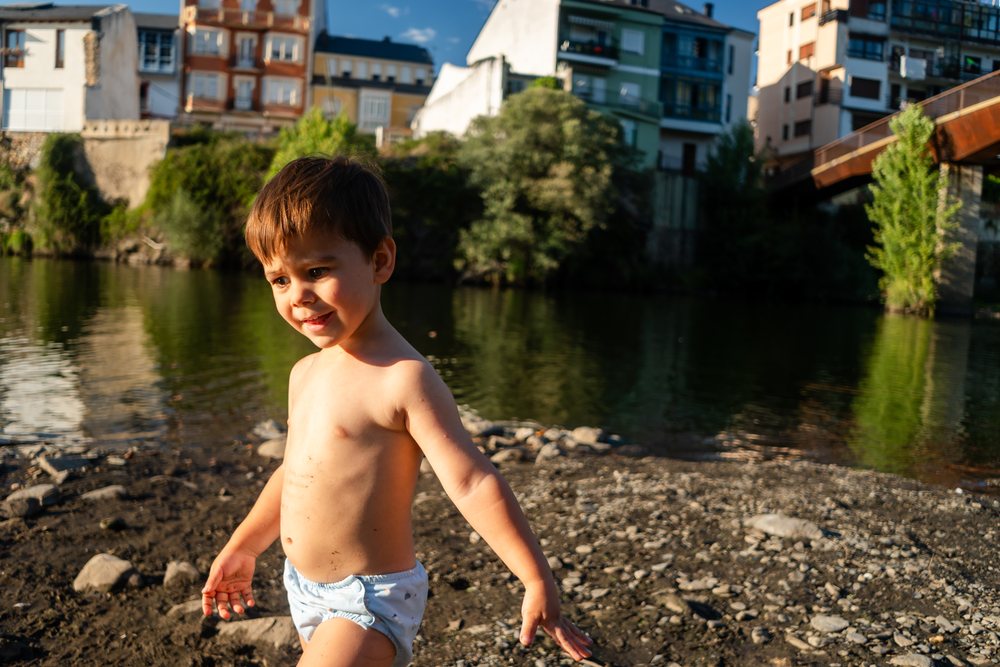 A toddler white boy plays by the riverbank with town buildings behind