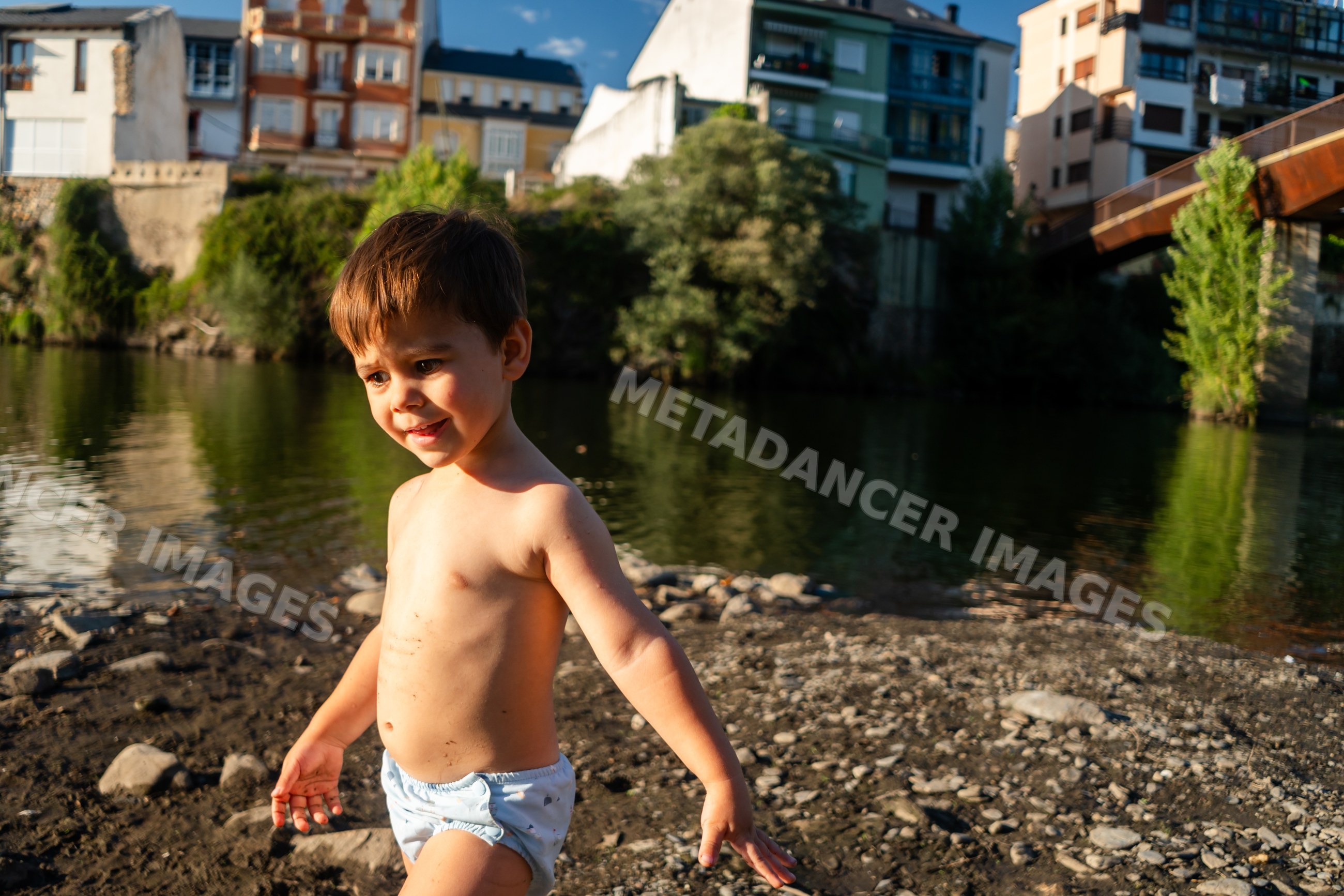 A toddler white boy plays by the riverbank with town buildings behind