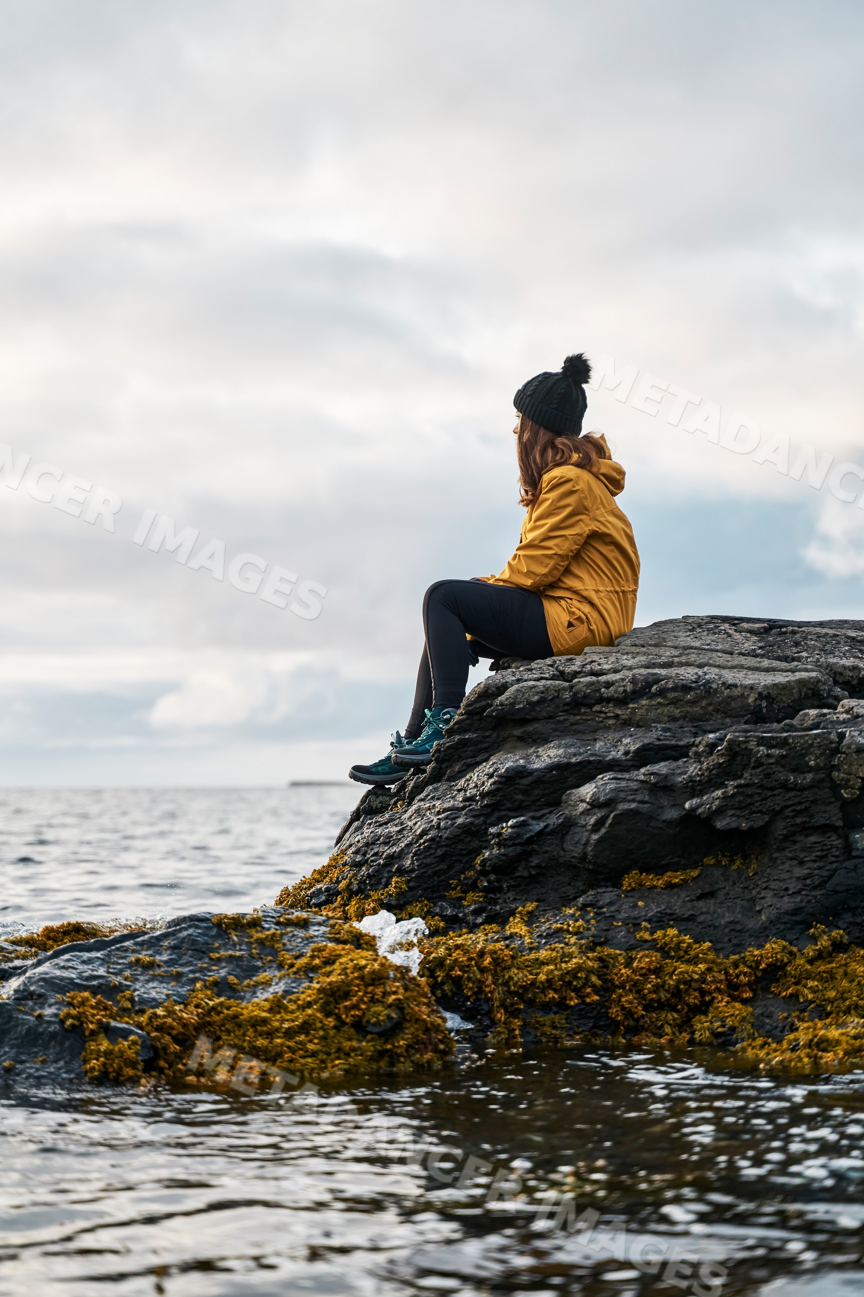 A white woman sits on a rocky coast by the ocean