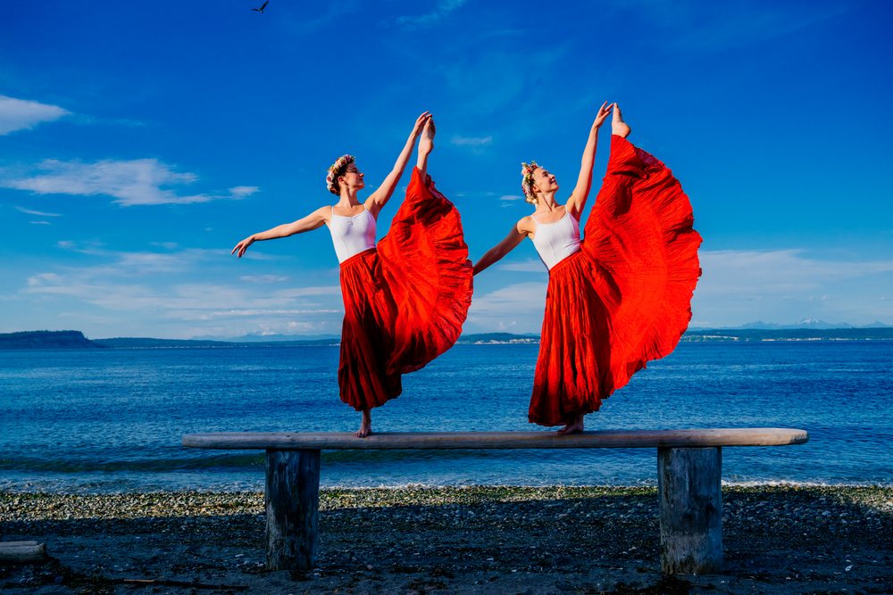 Two white women performing partner acrobatics yoga poses by the water