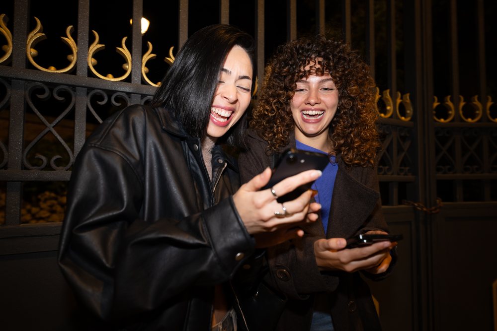 Two Hispanic and white women laughing and sharing smartphones outdoors