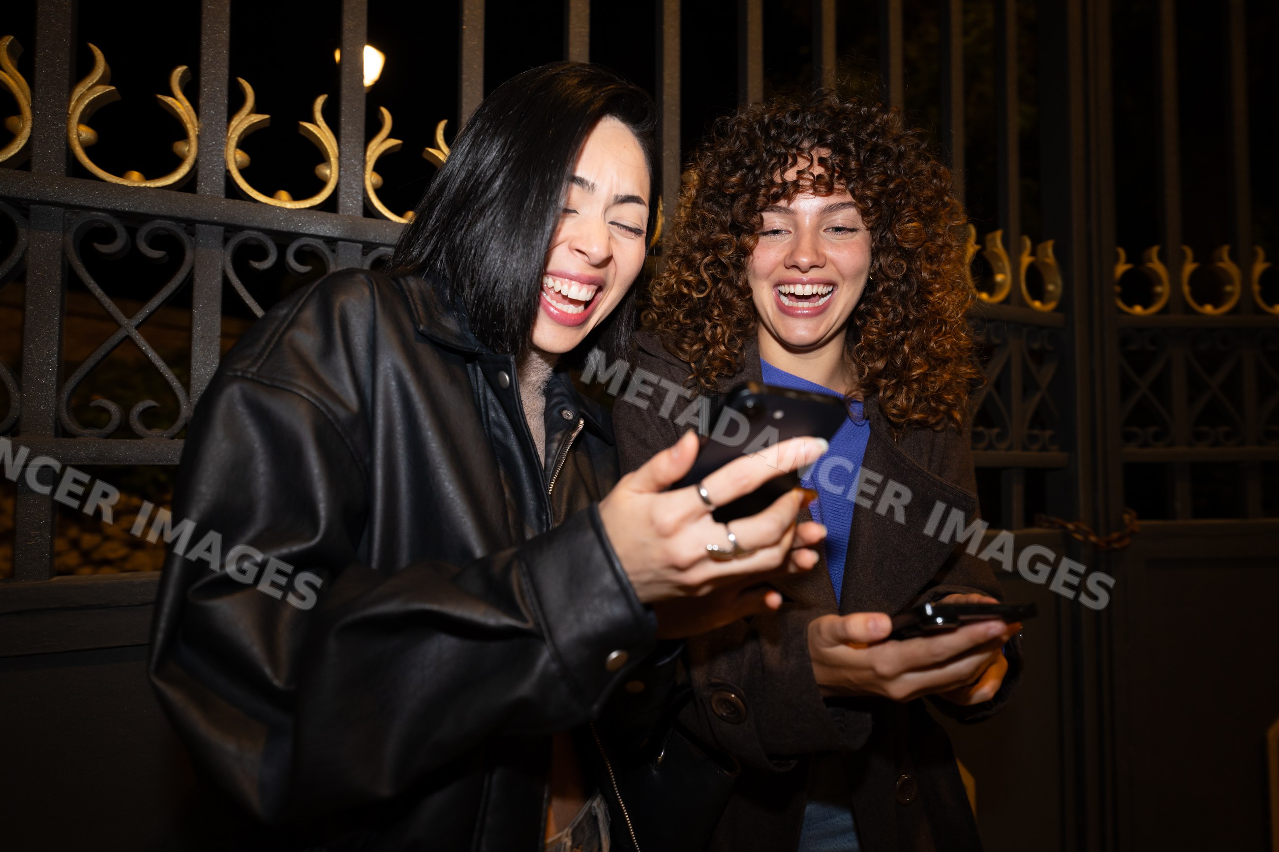 Two Hispanic and white women laughing and sharing smartphones outdoors