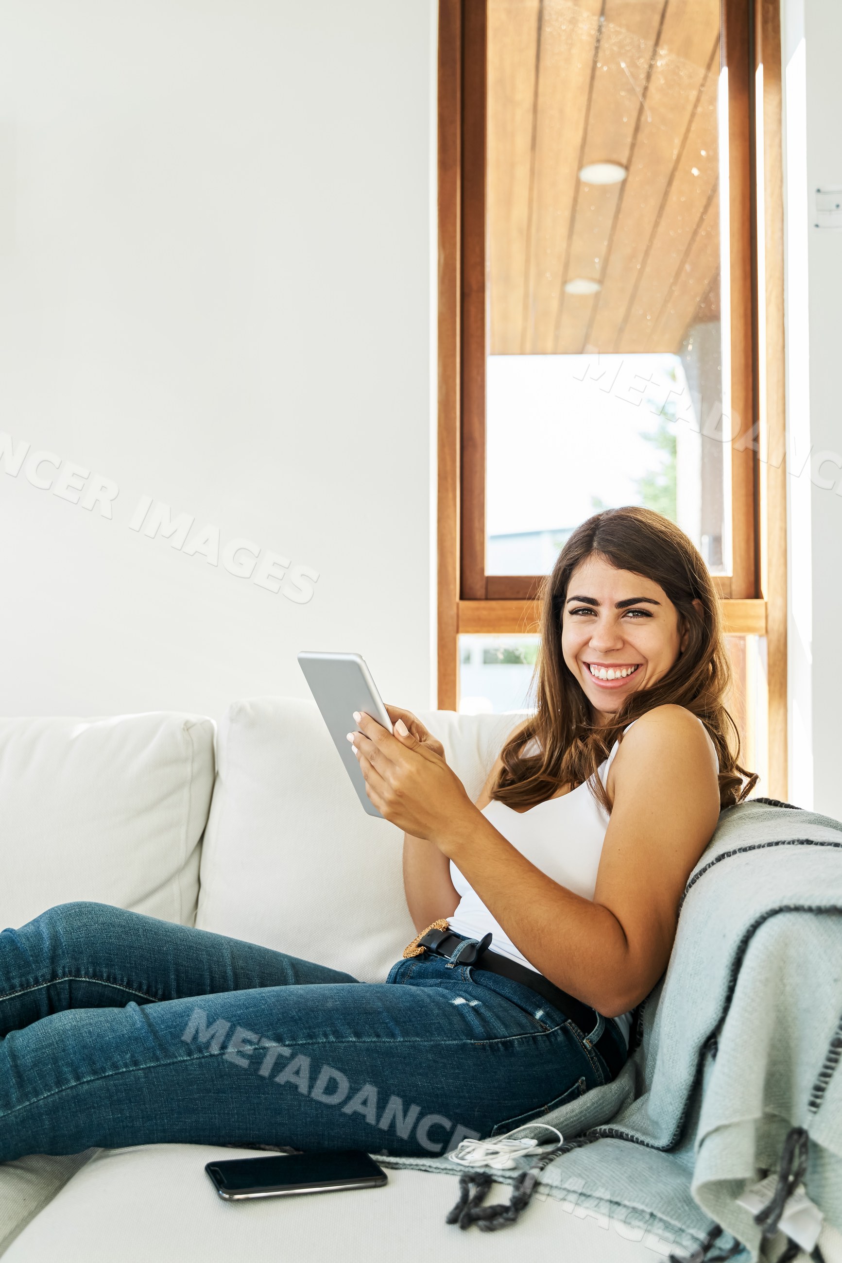 A smiling white woman relaxes on a sofa using a tablet