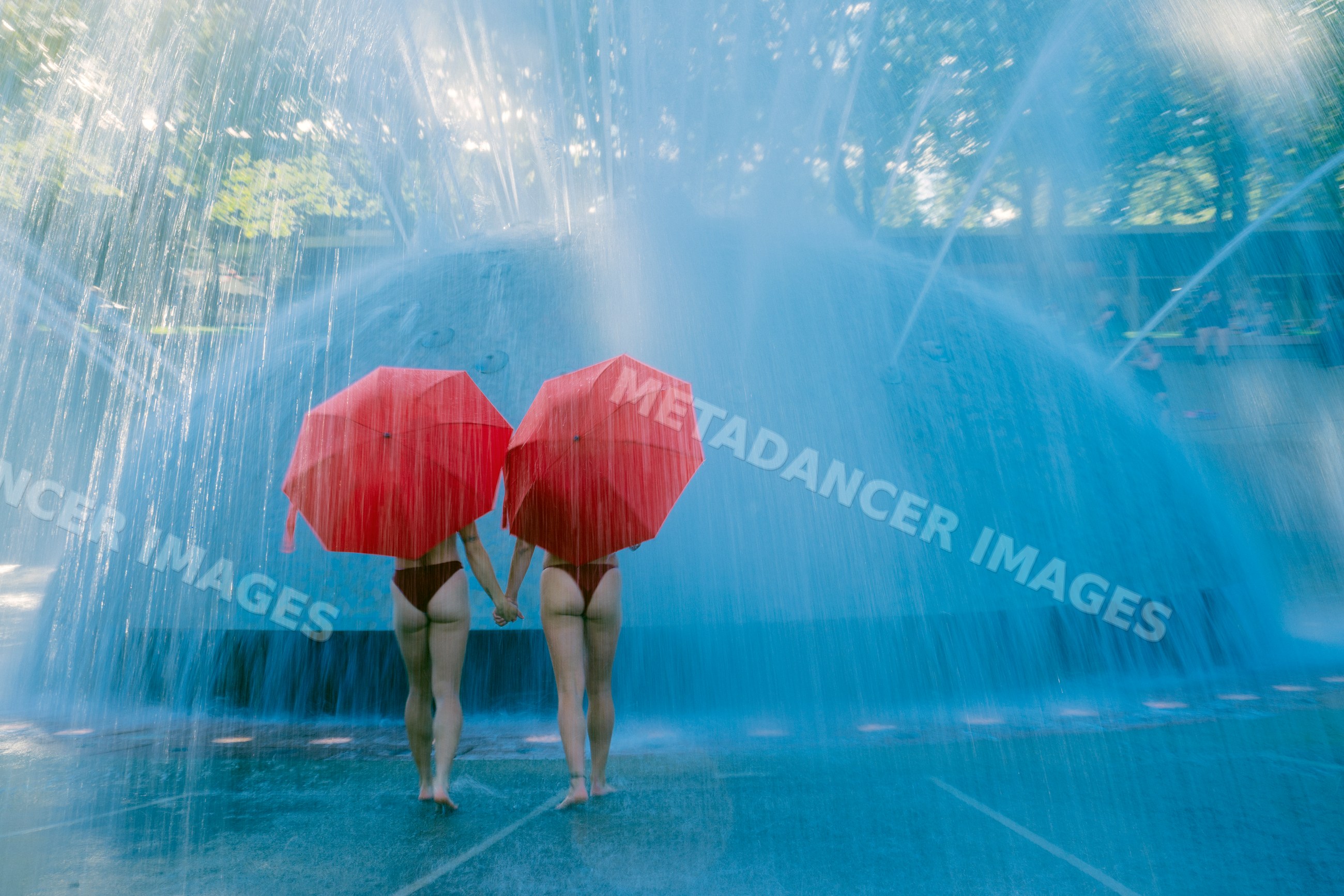 Two white women holding umbrellas walking through a water fountain