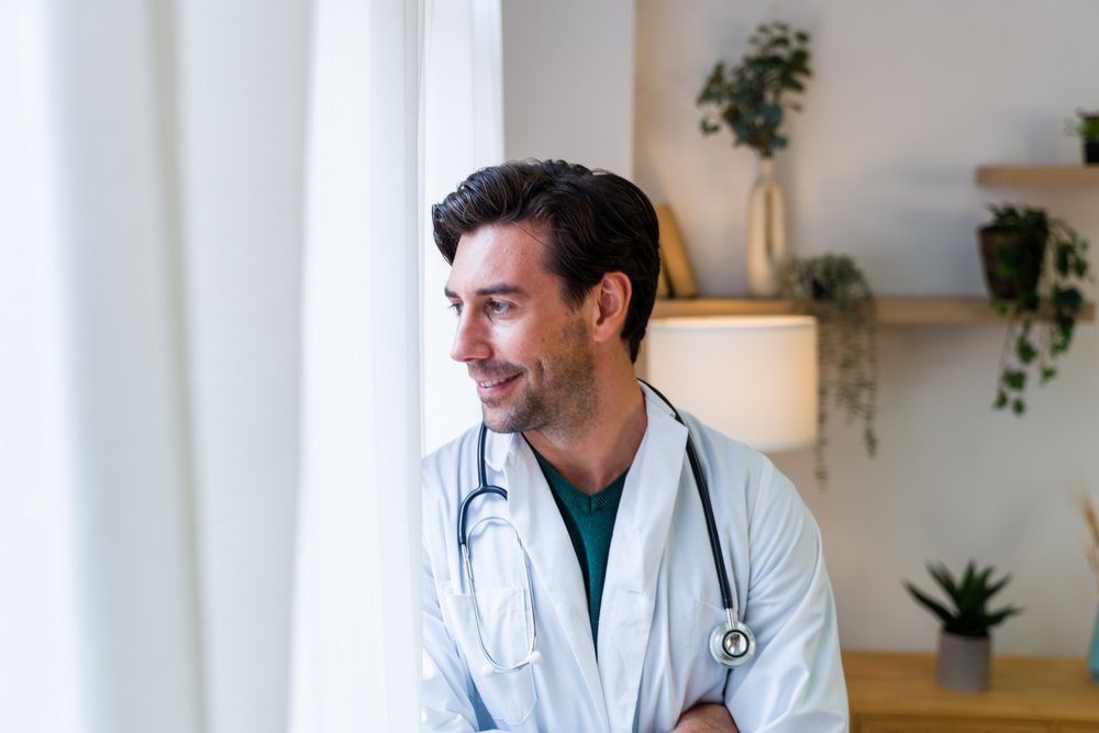 Male doctor in white coat looking out window inside medical office
