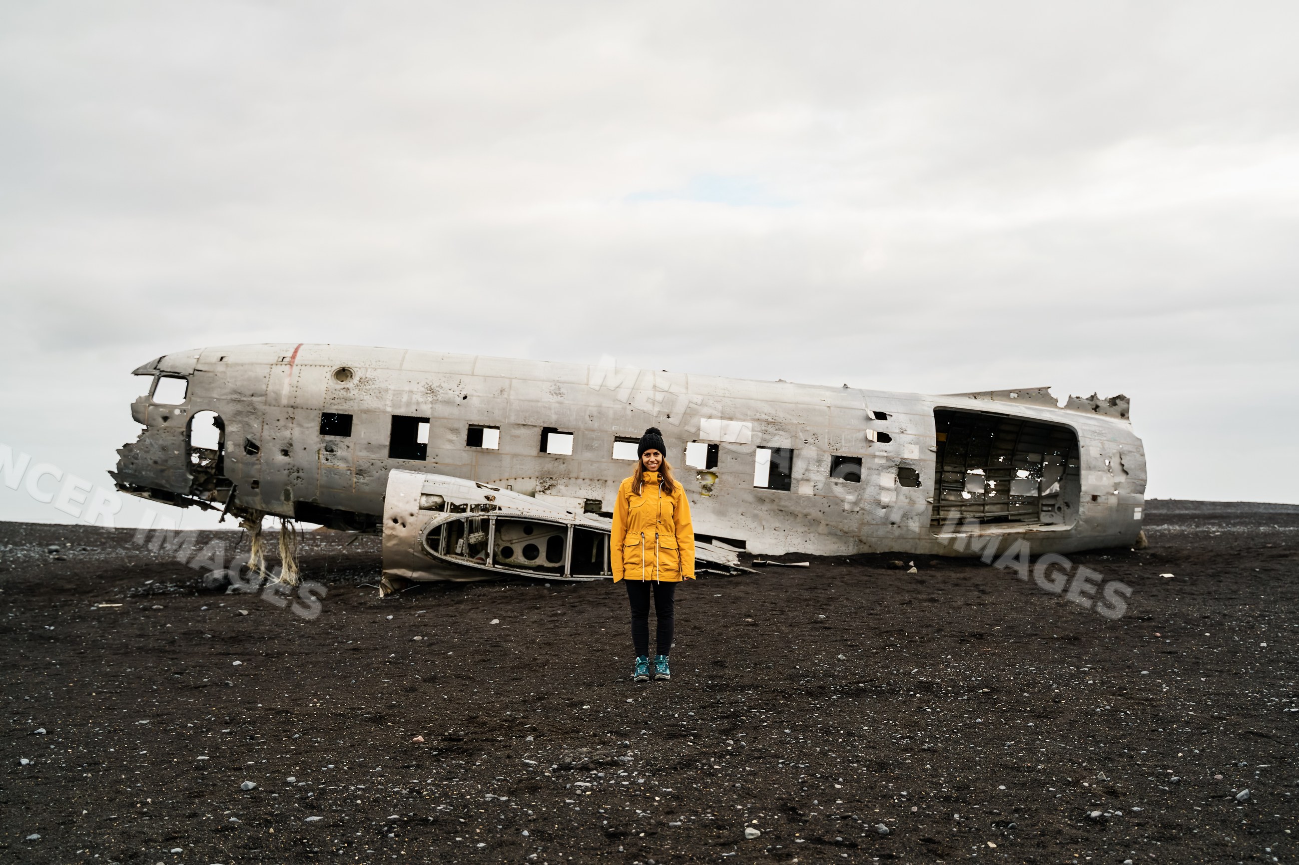 A white woman stands before an airplane wreck on black volcanic soil