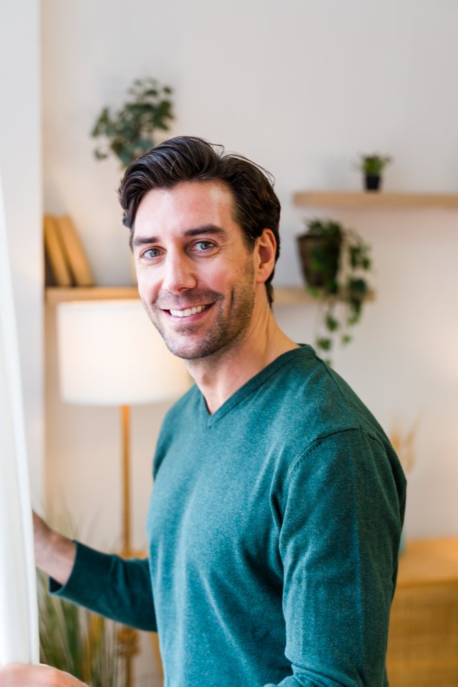 White man smiling indoors while holding curtain in a living room