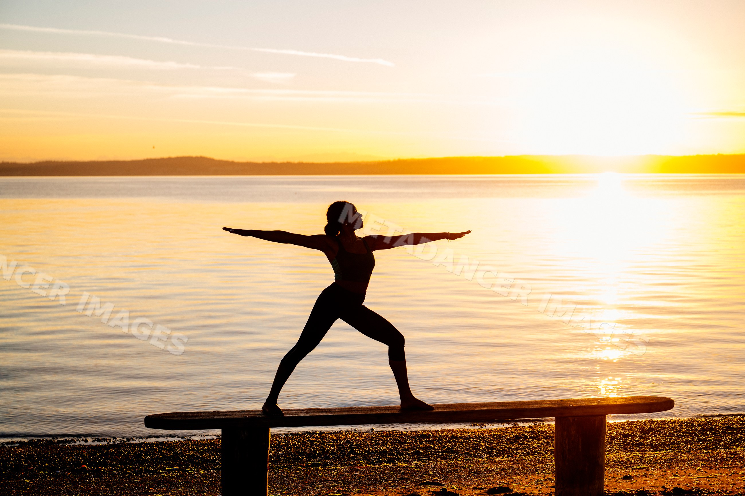 Hispanic woman practicing yoga warrior pose on bench at beach sunset