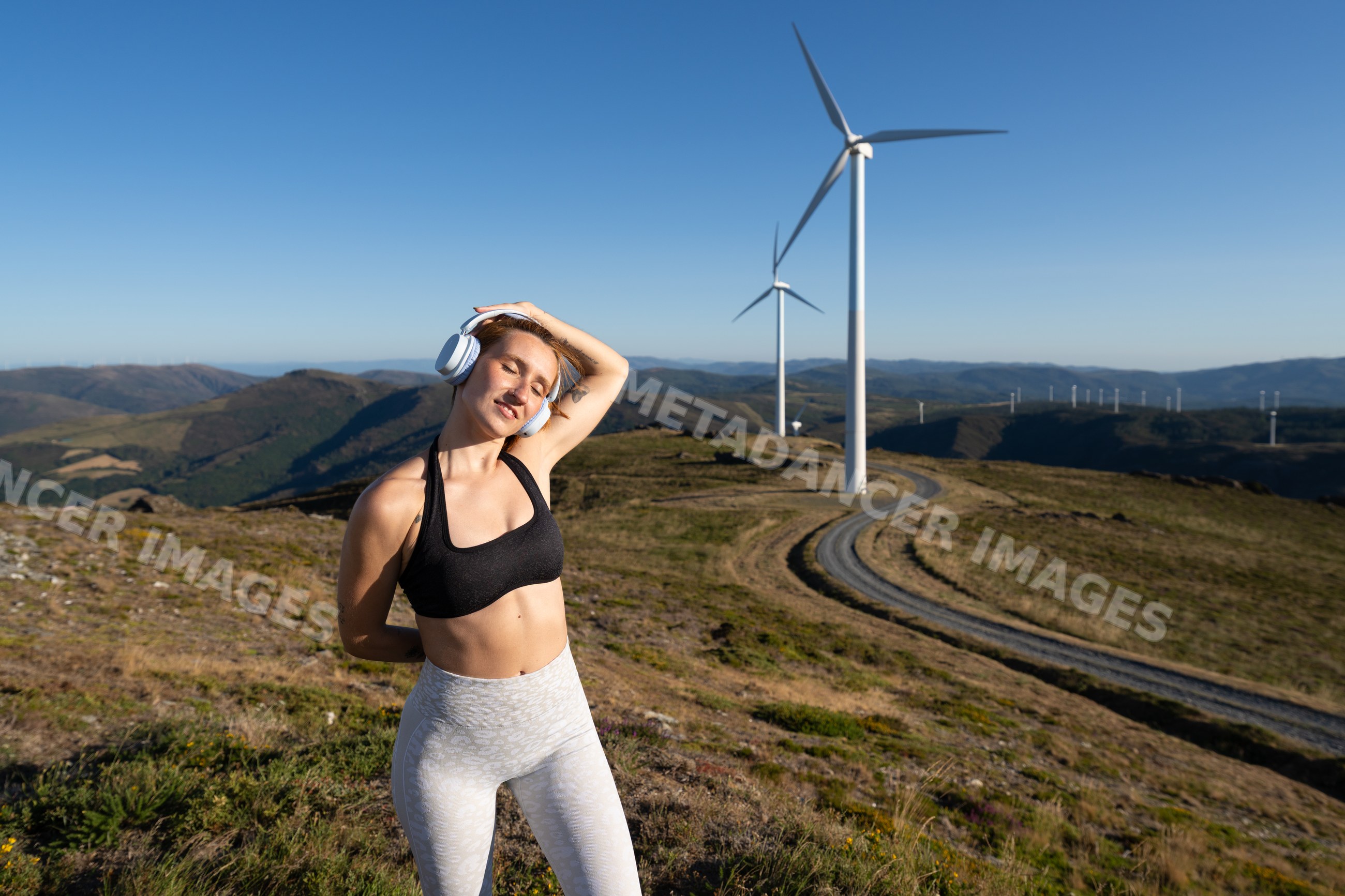 A white woman stretches near wind turbines on a sunny day in Galicia