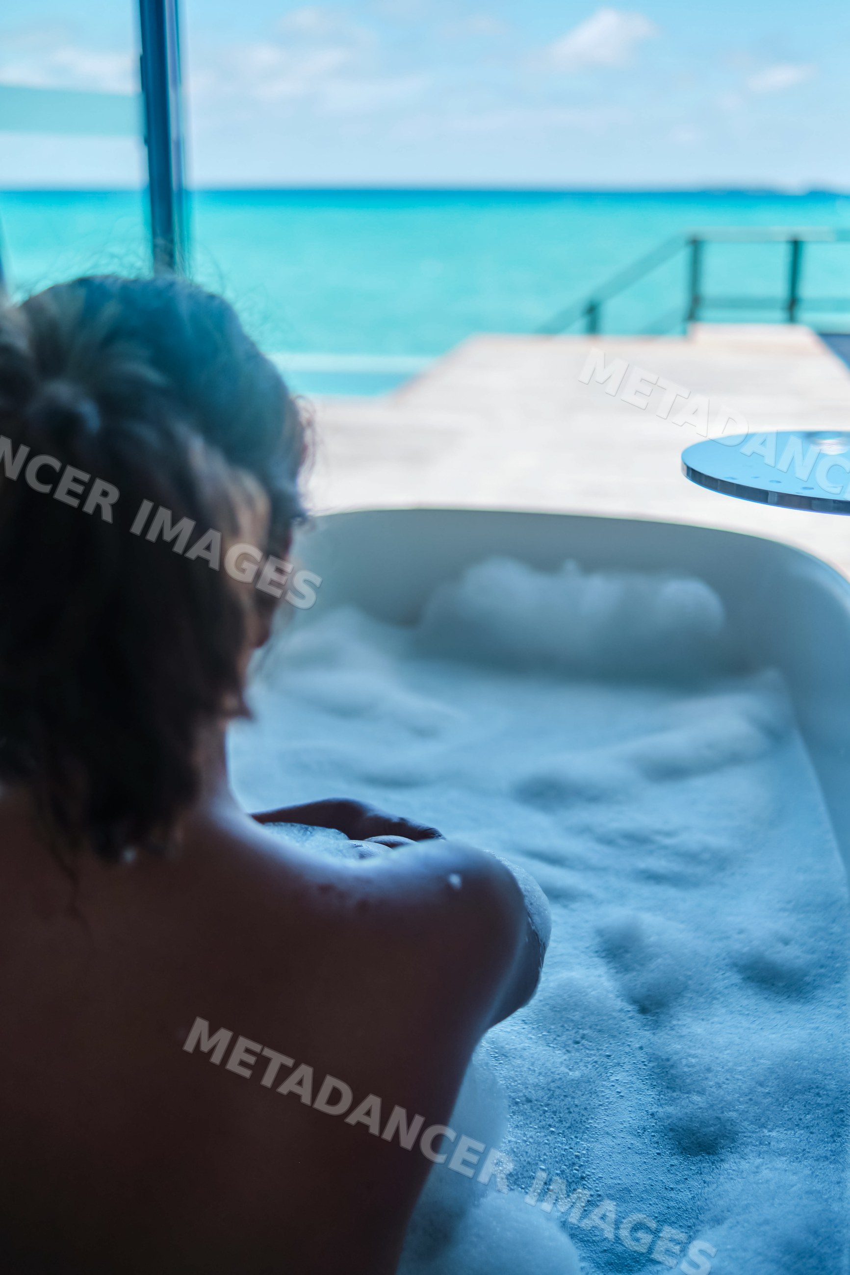 A white woman relaxes in a bubble bath overlooking the ocean