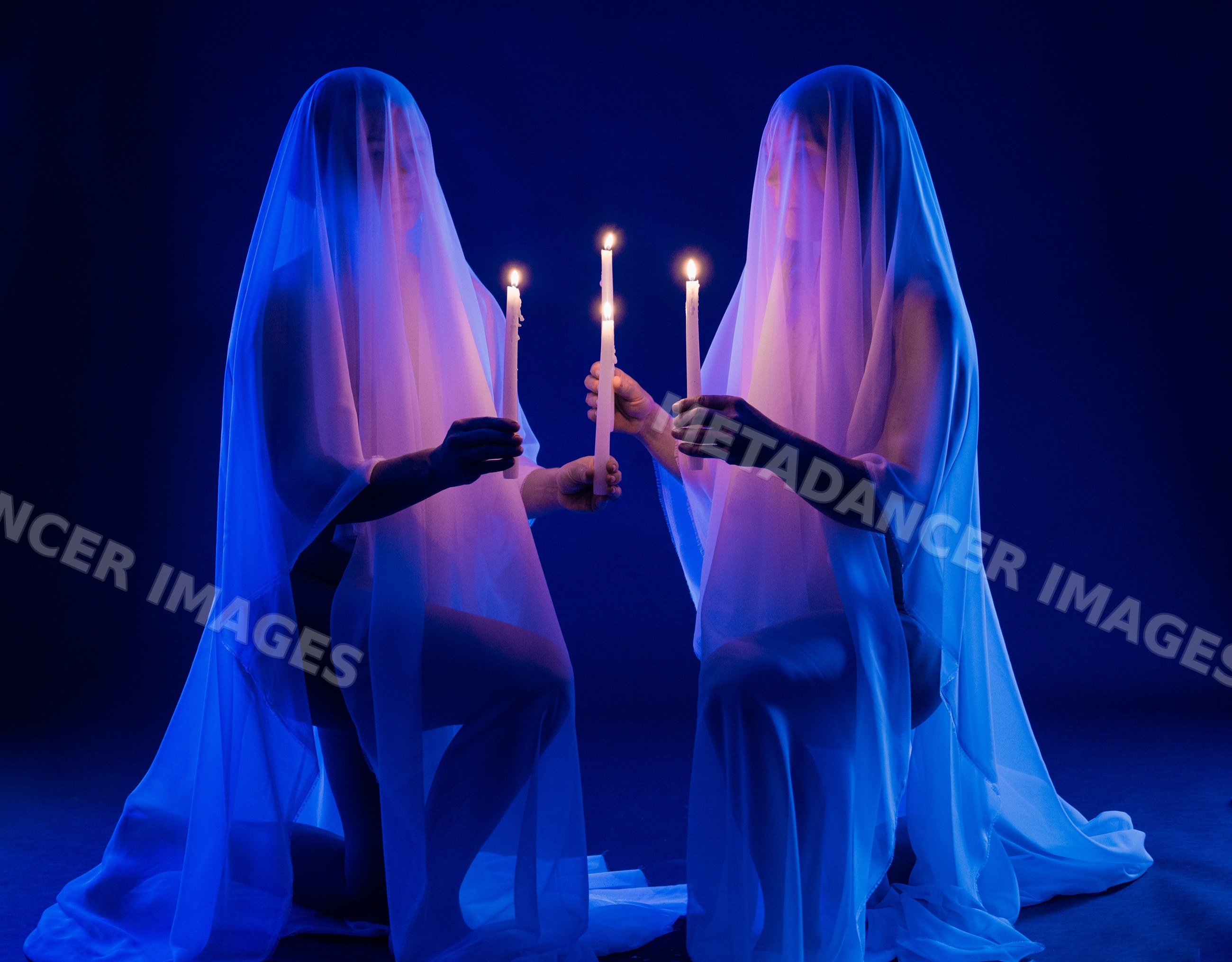 Two veiled kneeling figures hold candles in blue-lit dark studio