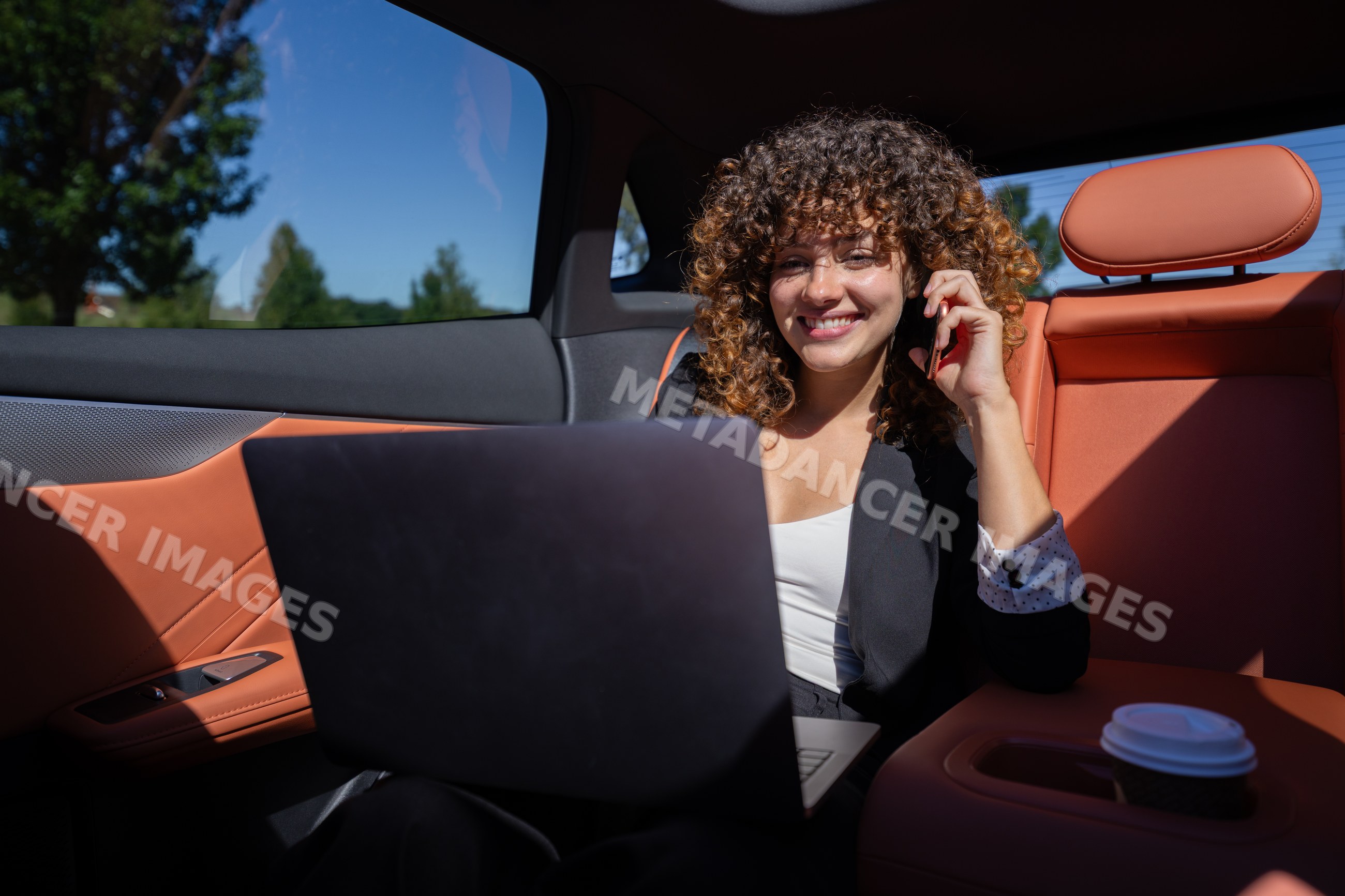 White woman working remotely inside car with laptop and phone