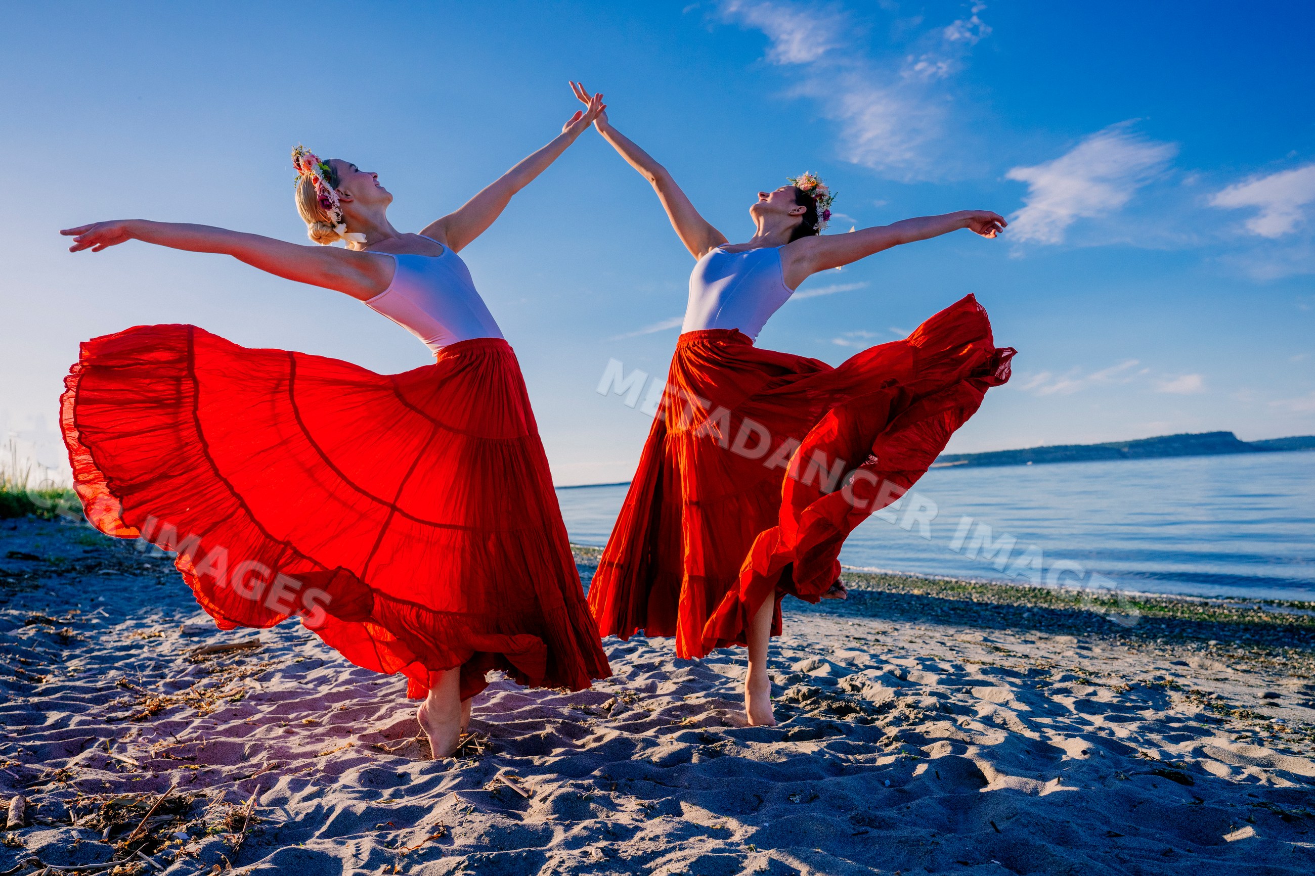 Two white women dancing gracefully on a beach with flowing red skirts