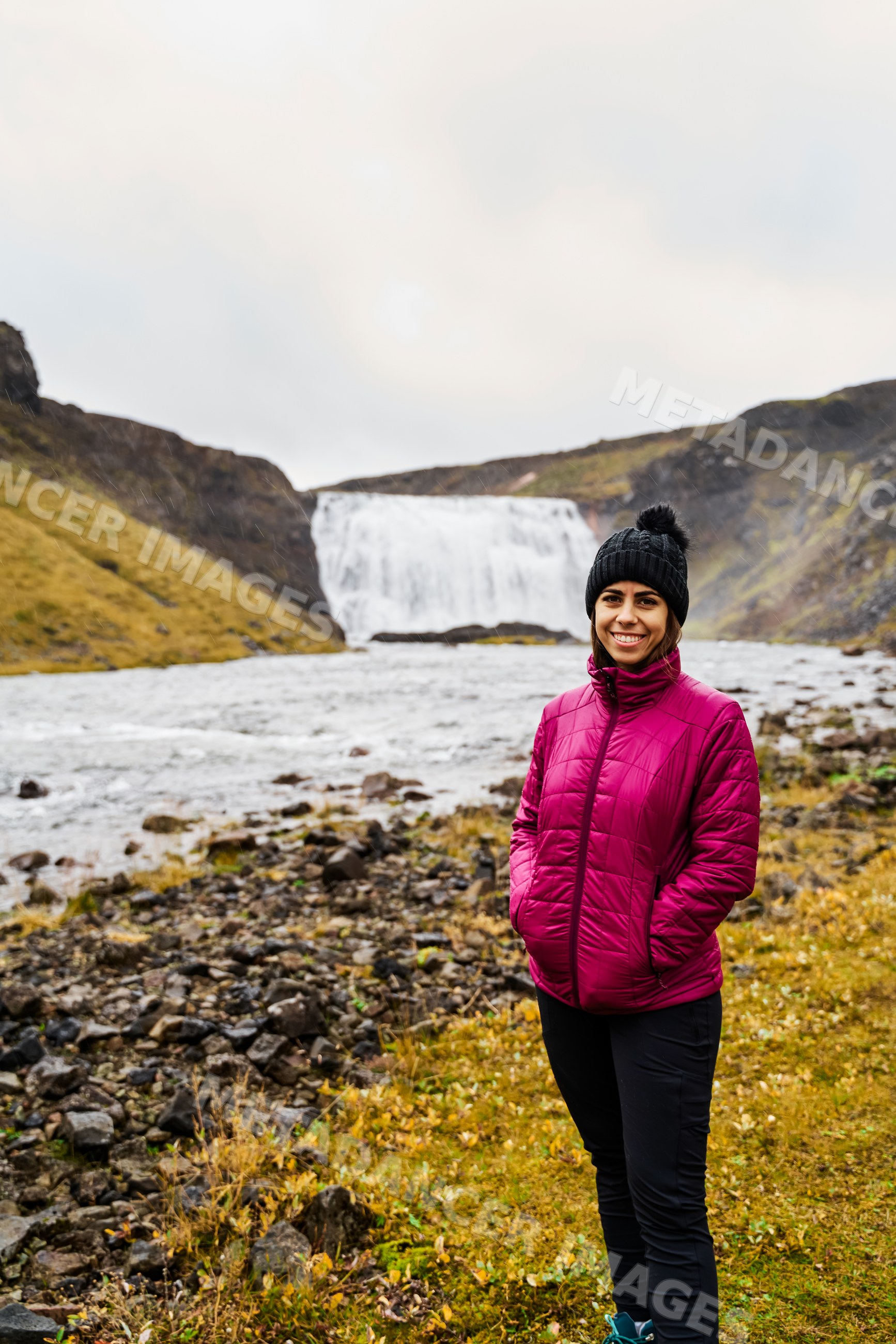 A white woman stands smiling by a river with a waterfall