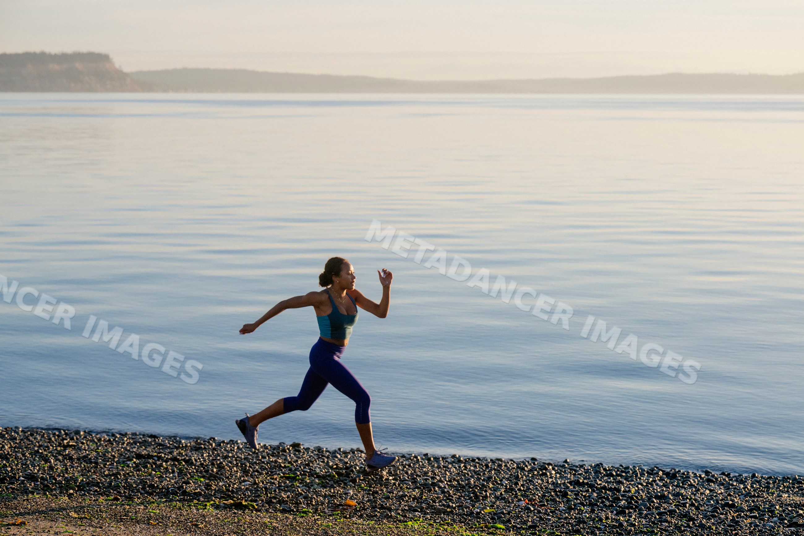 Hispanic woman running on rocky beach shoreline with calm water
