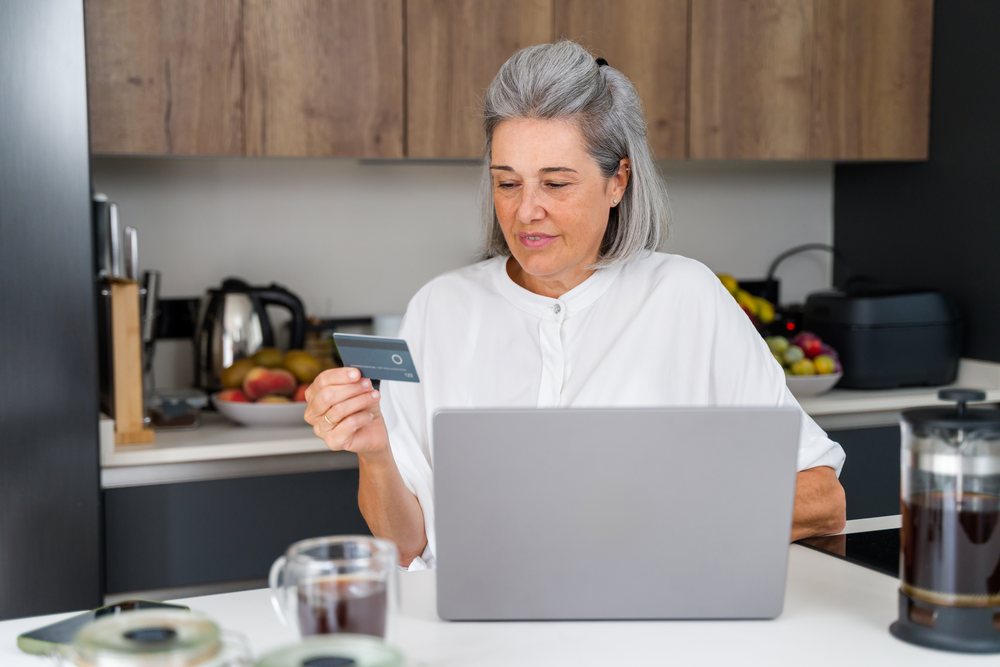 Senior white woman making online payment on laptop in kitchen in Spain