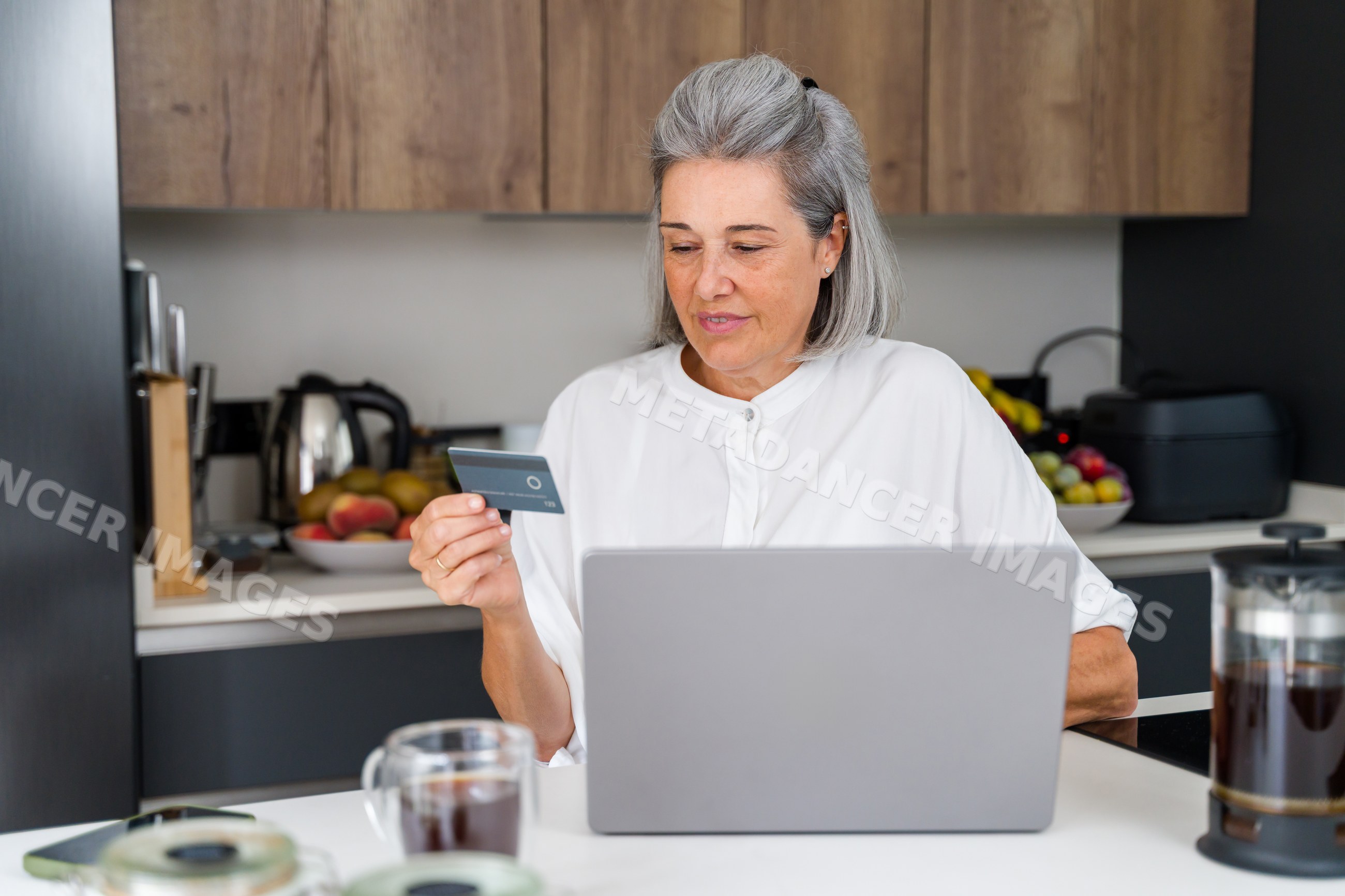 Senior white woman making online payment on laptop in kitchen in Spain