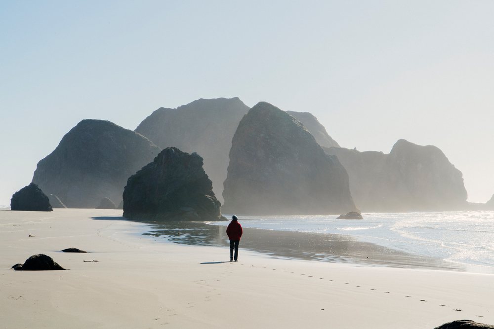 White person walking on beach near large coastal rocks and ocean waves