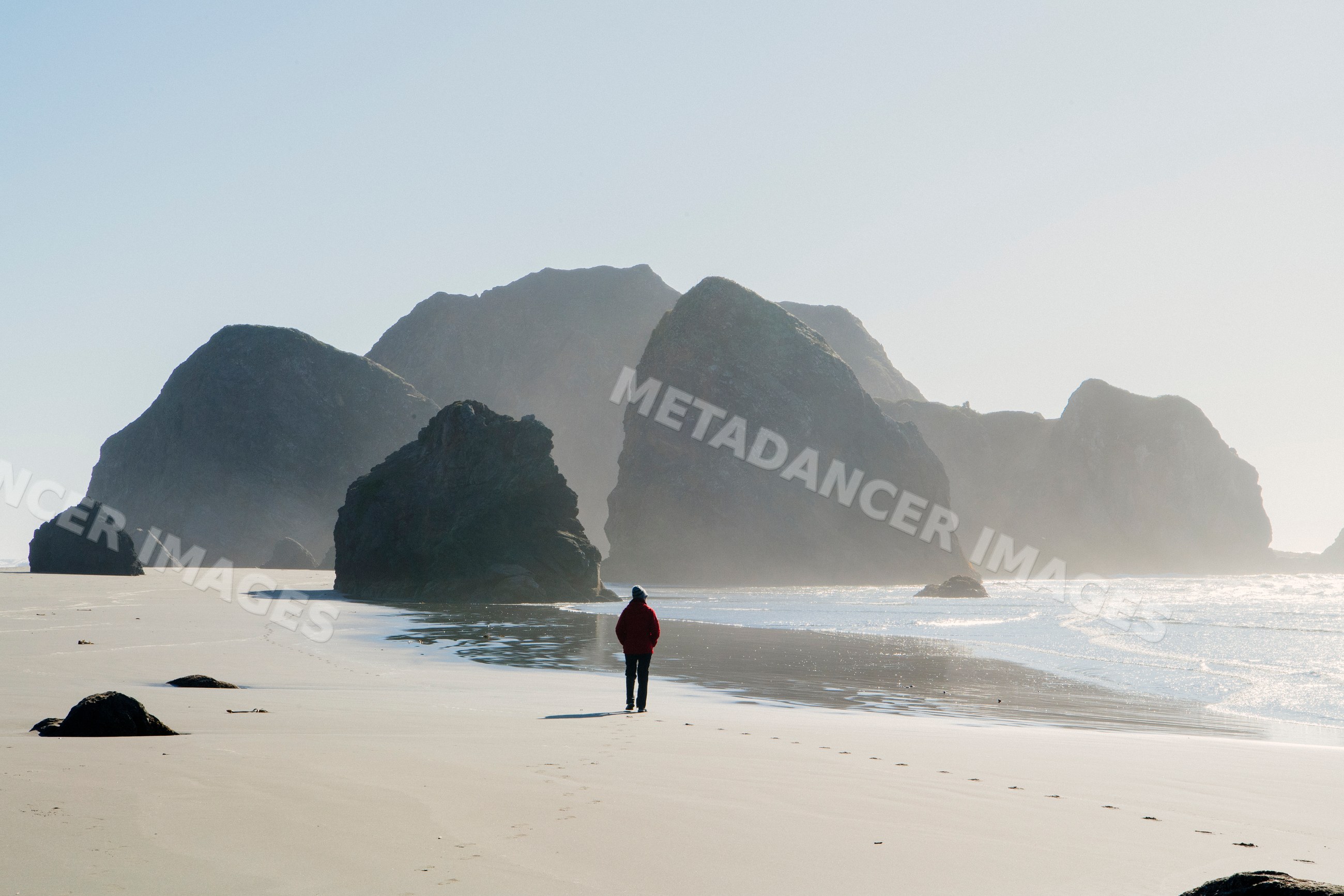 White person walking on beach near large coastal rocks and ocean waves