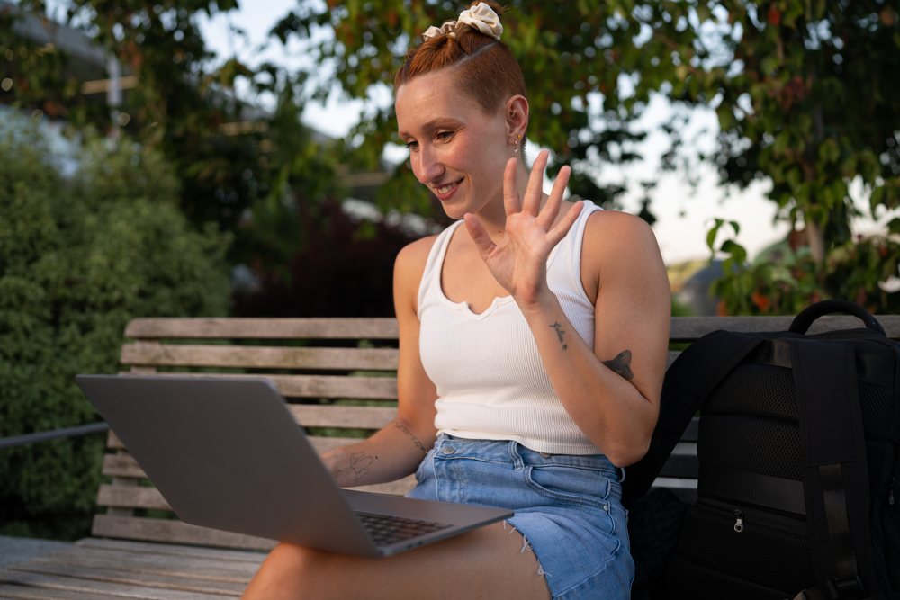 Young white woman waves during a video call on a park bench