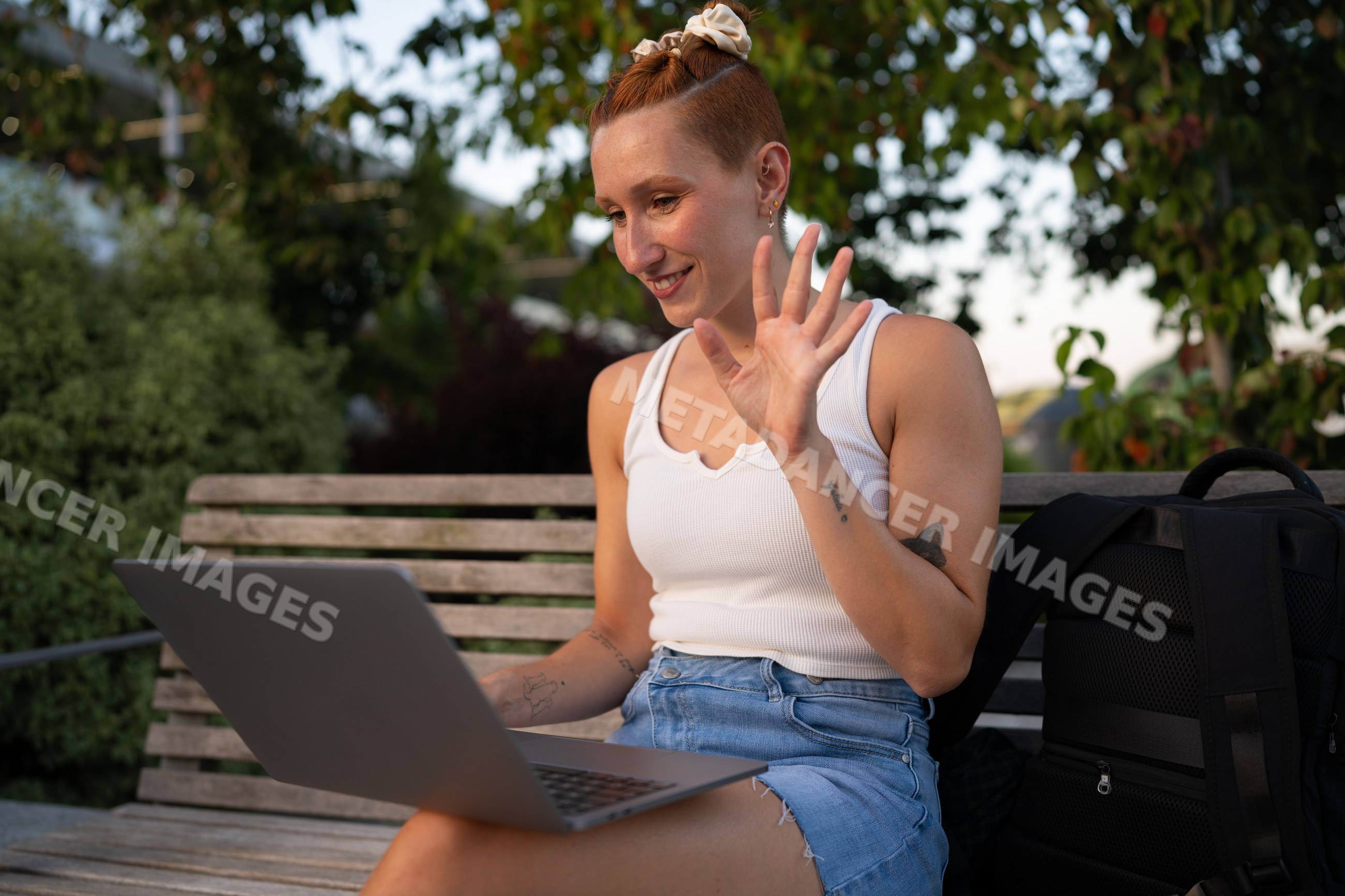 Young white woman waves during a video call on a park bench