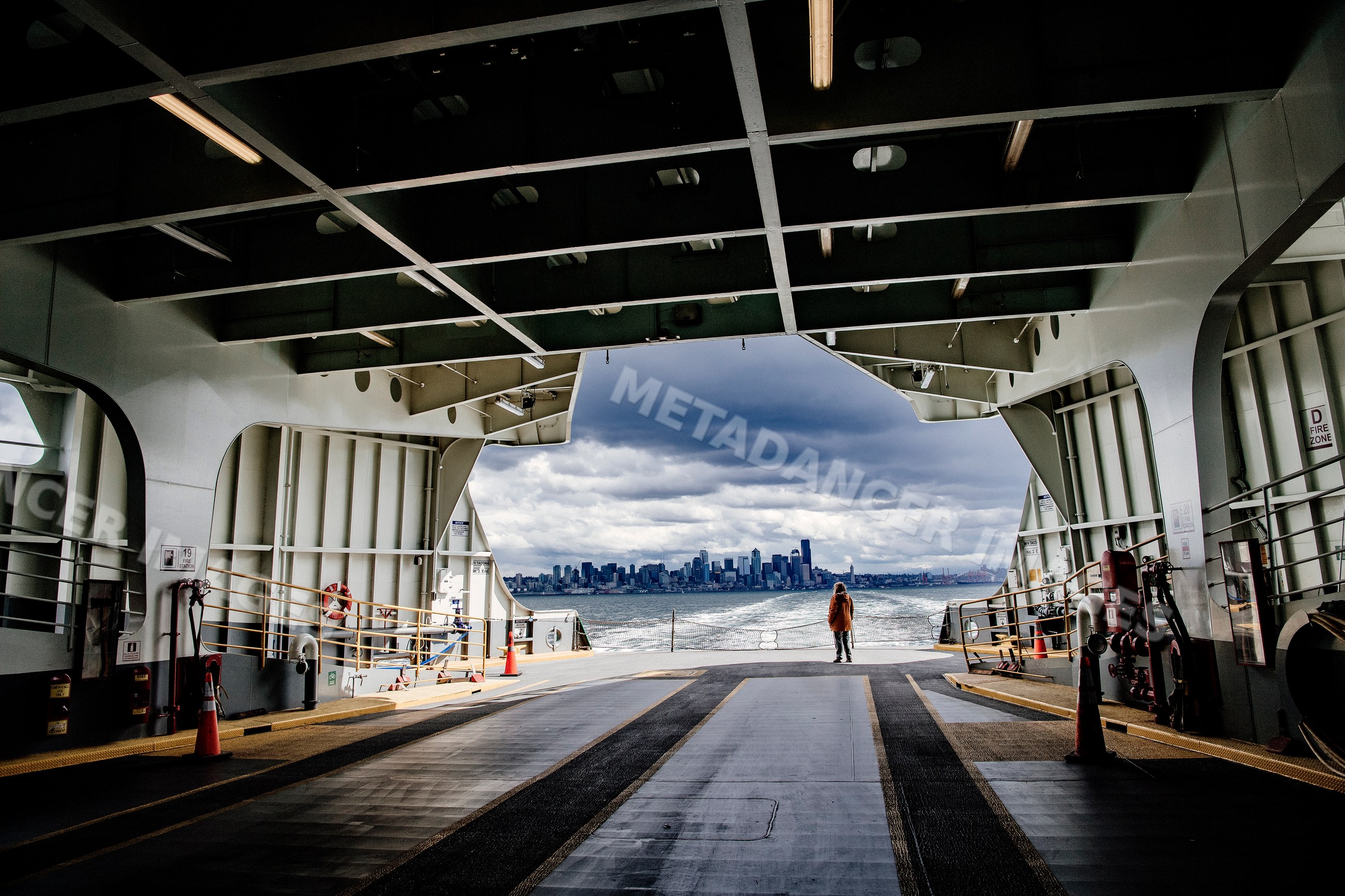 A person stands on a ferry deck viewing a city skyline on an overcast
