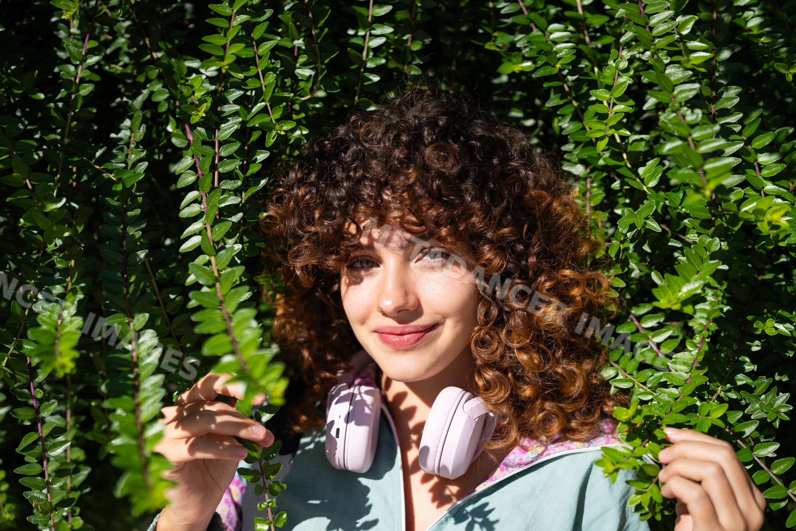 White woman with curly hair wearing headphones among green foliage