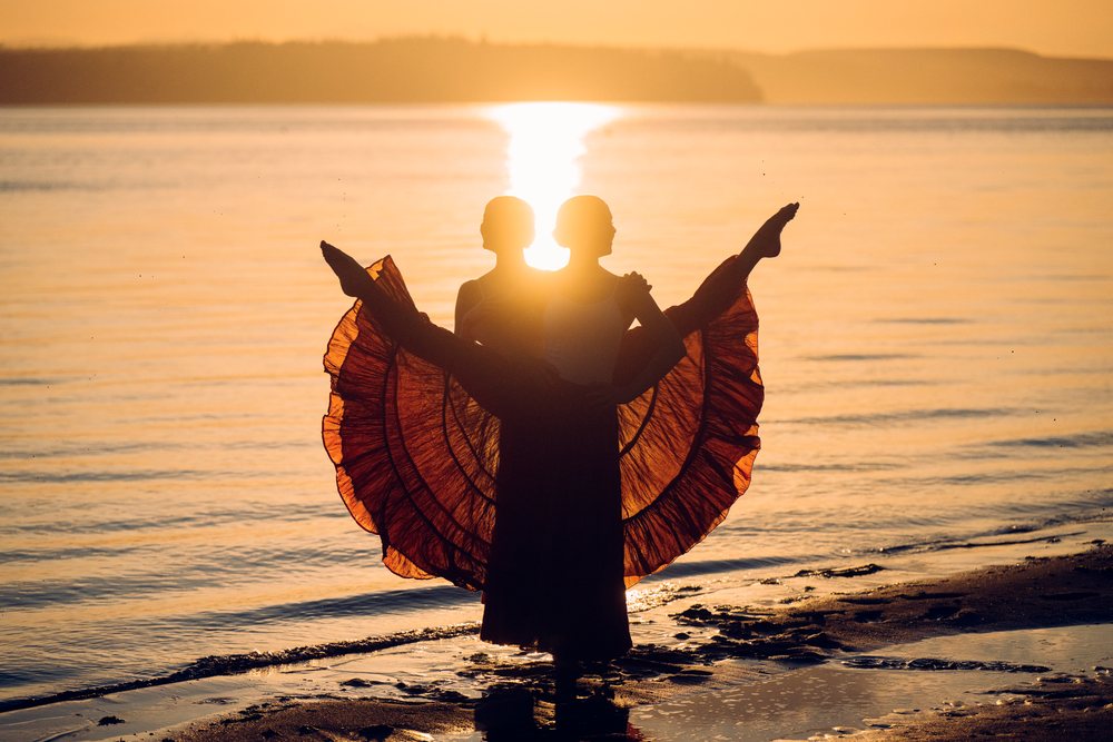 Two white women performing partner acrobatics on a beach at sunset